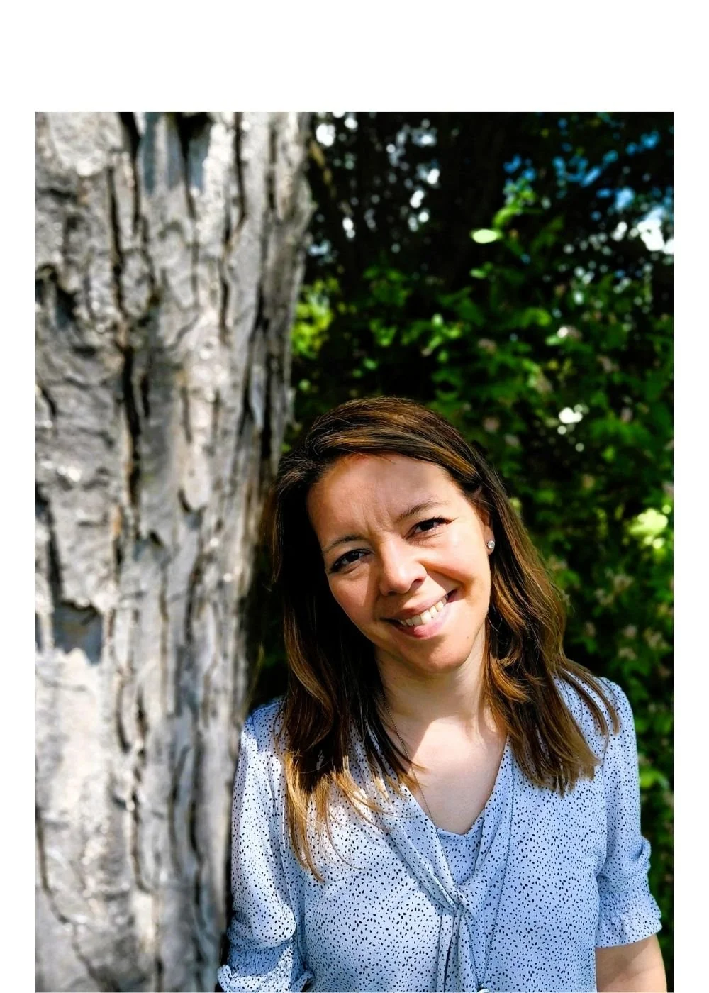 A woman with brown hair and a light blue, polka-dotted blouse smiling outdoors near a large tree with green foliage in the background.