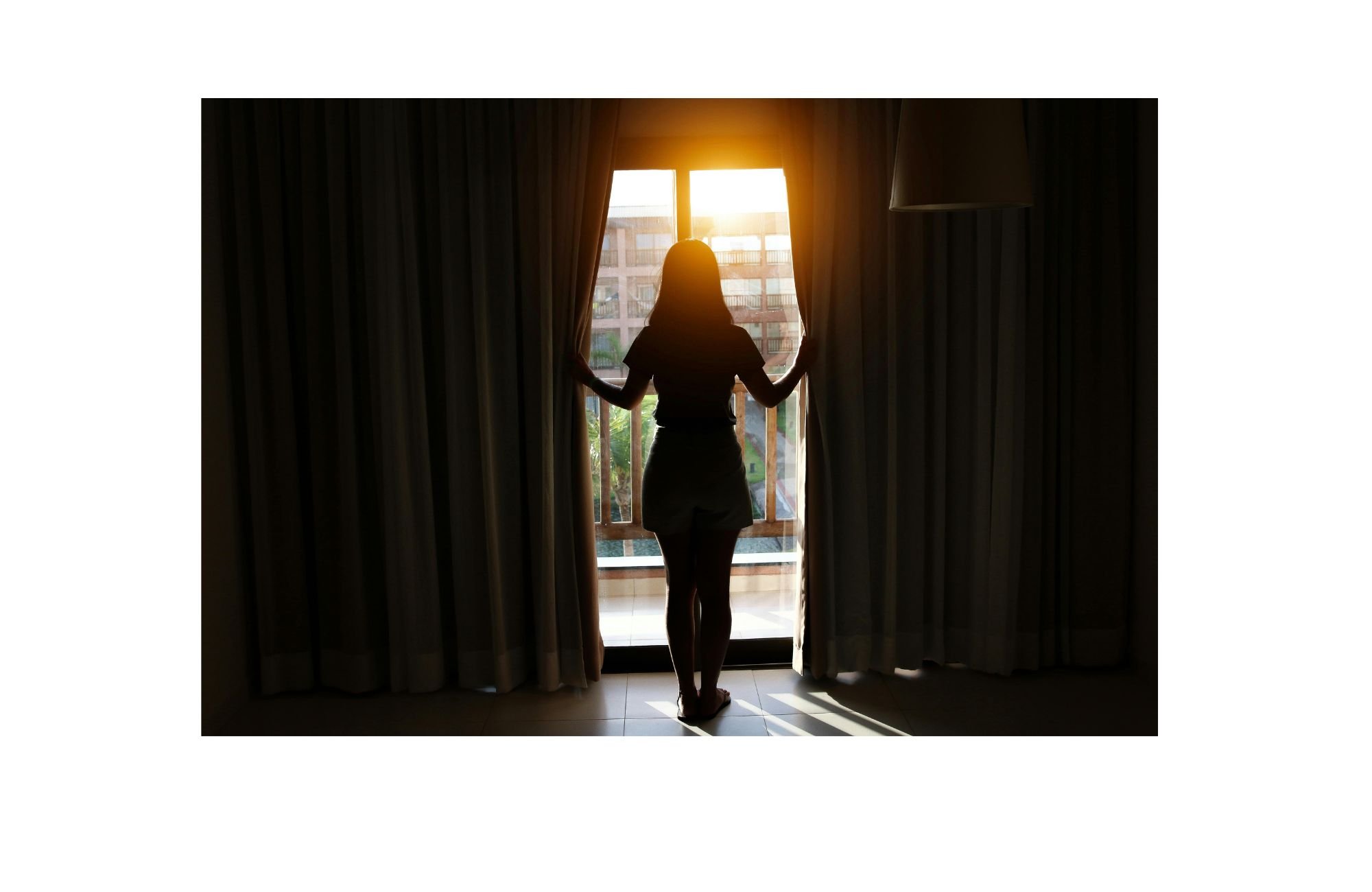 A woman standing by a window, opening curtains to view the outdoors during sunrise or sunset.