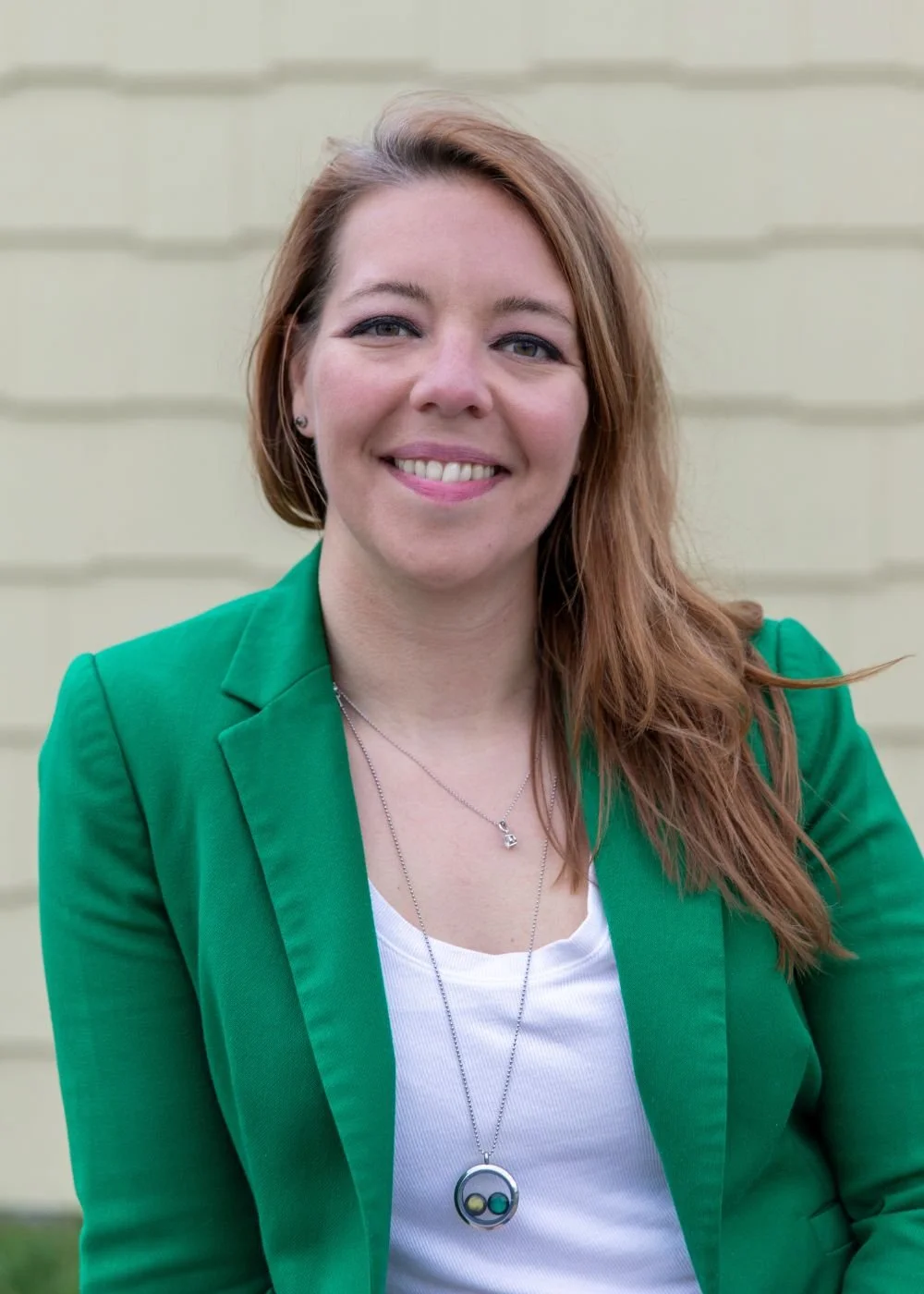 Portrait of a woman with red hair wearing a green blazer and a necklace, smiling at the camera against a pale yellow background.