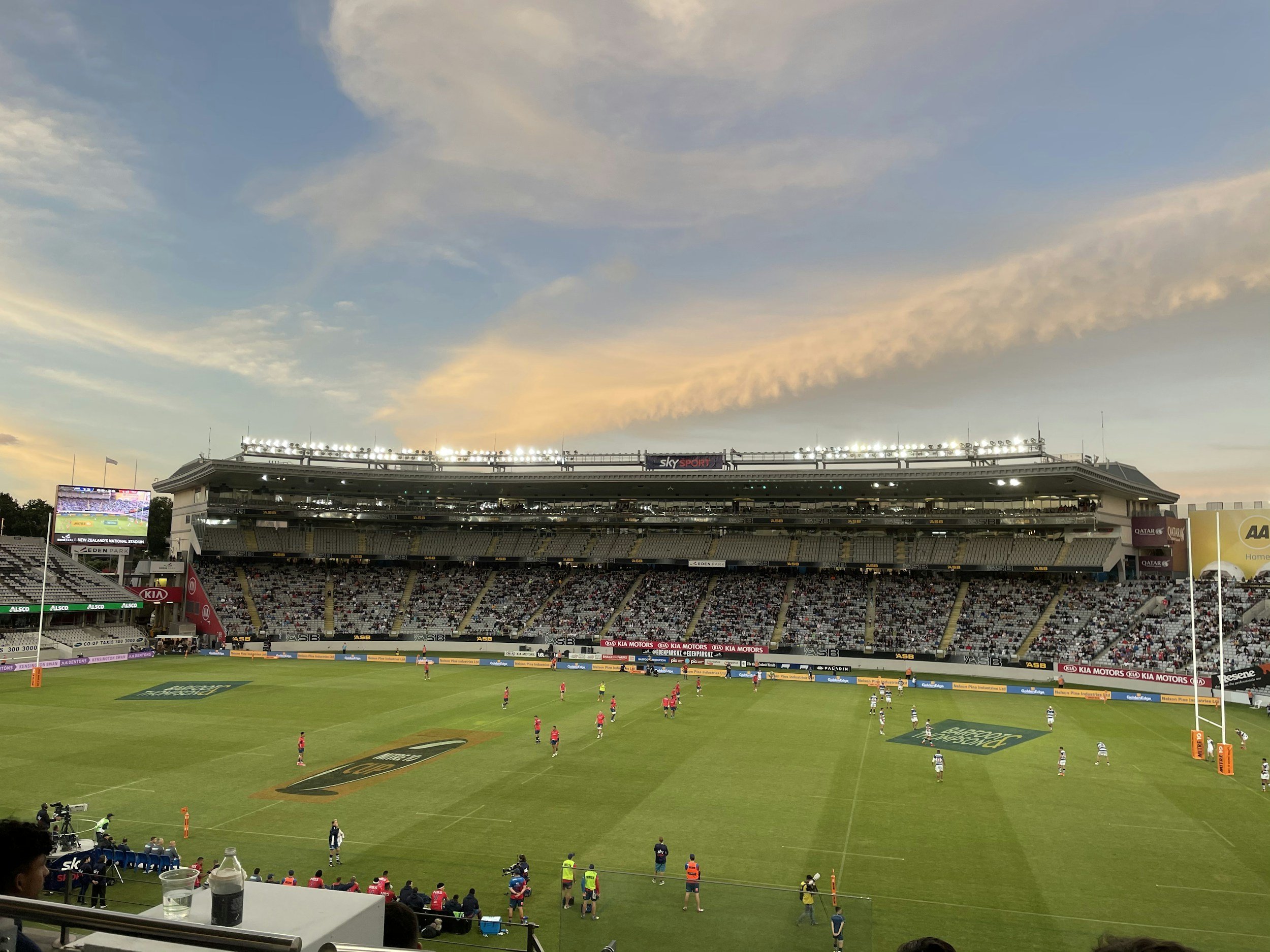 A rugby stadium filled with spectators, players on the field warming up, and sky with light clouds before sunset.