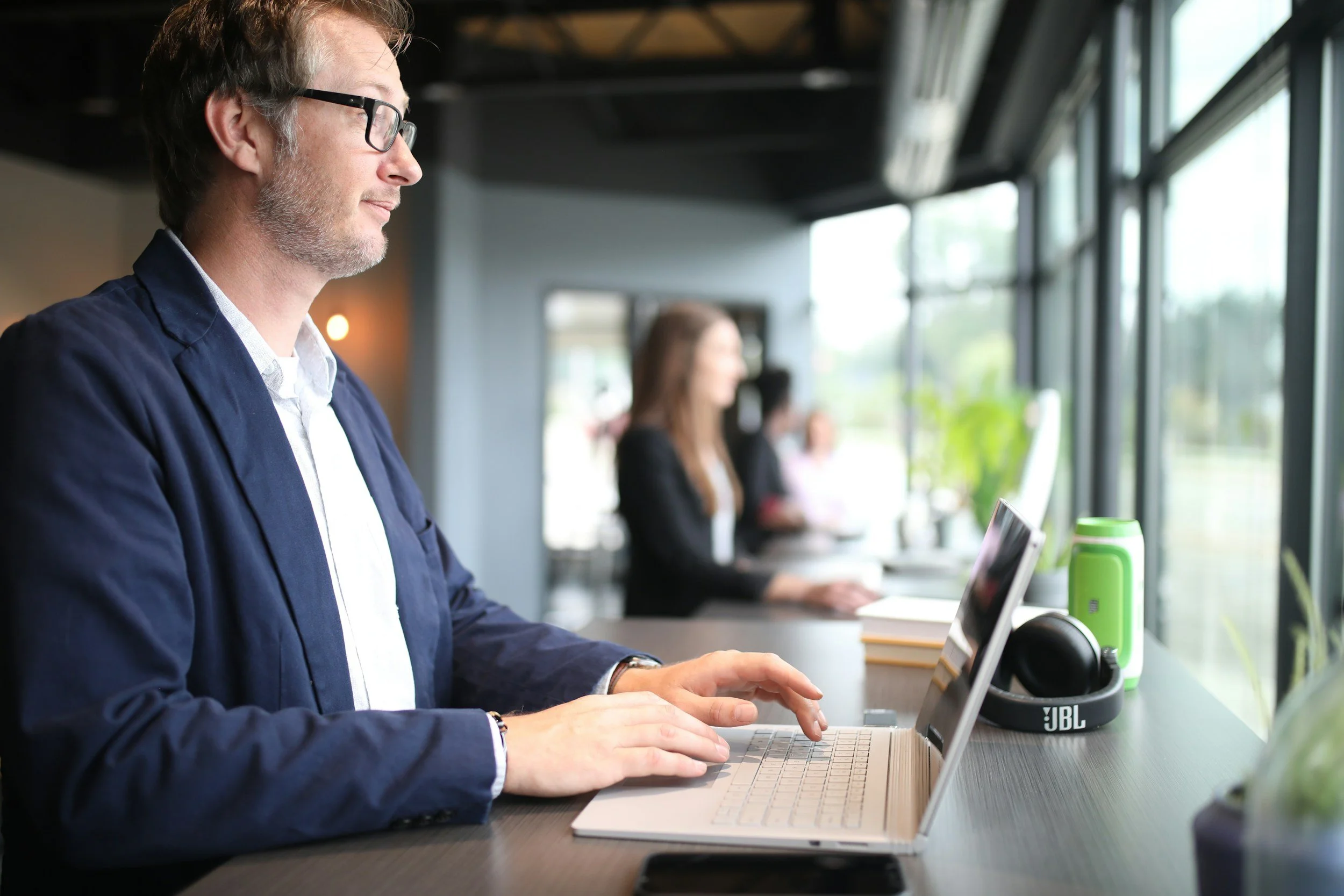 A man with glasses sitting at a desk using a laptop in a bright room with large windows, with a woman in the background also working on a laptop.