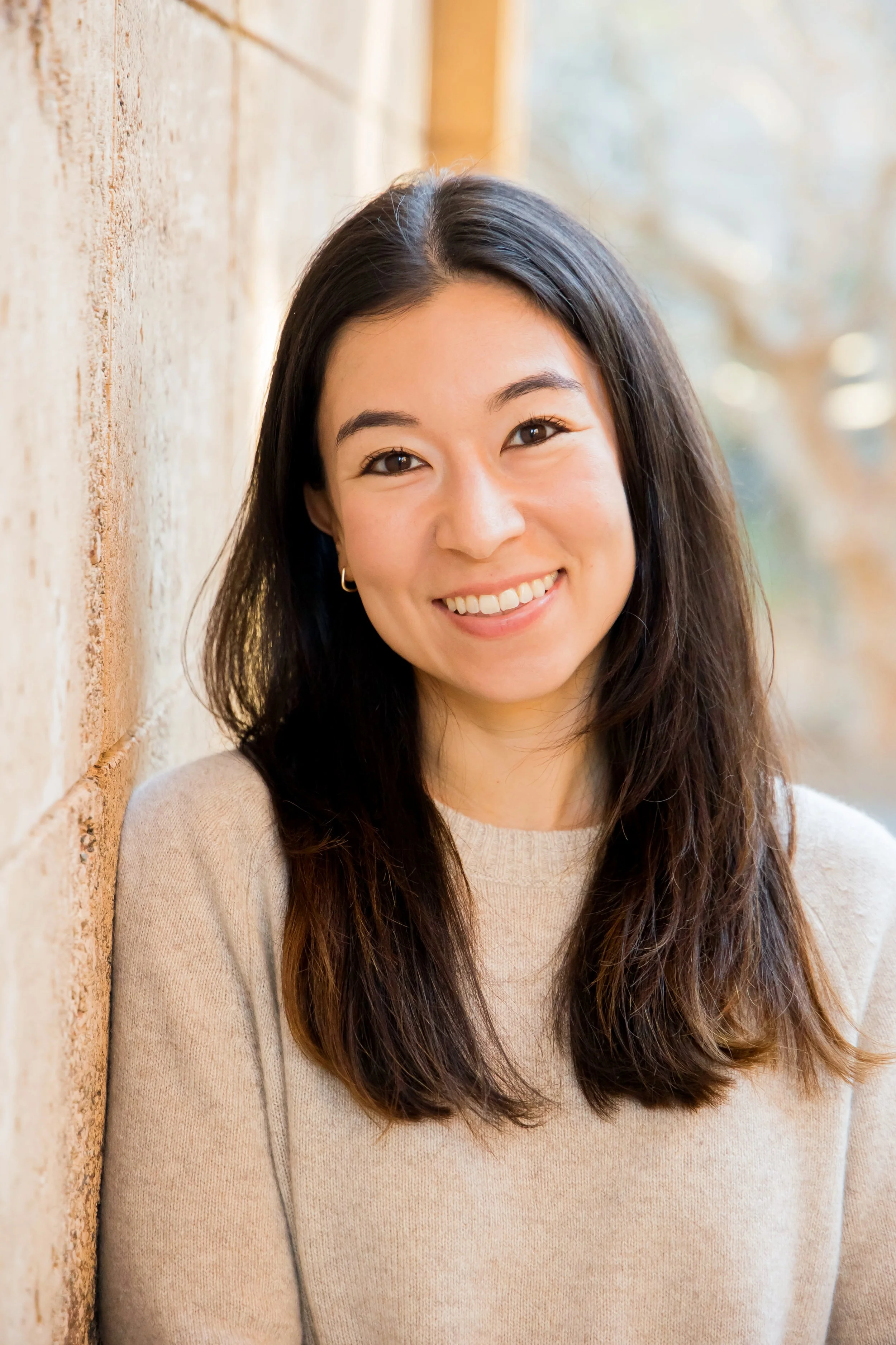 Dr. Emily Hunt, a therapist with long brown hair and a beige sweater smiling, leaning against a brick wall outdoors with blurred trees in the background.