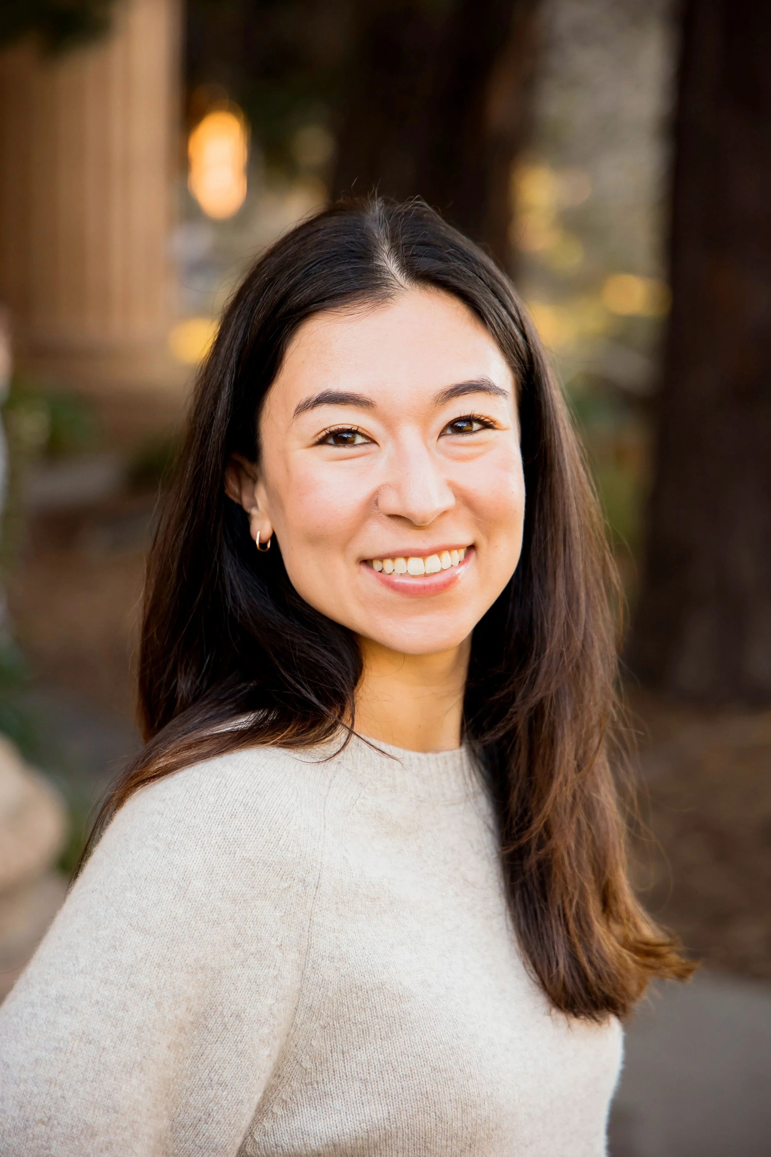 Dr. Emily Hunt, a therapist with long brown hair and a beige sweater, smiling outdoors with blurred trees and warm lighting in the background.
