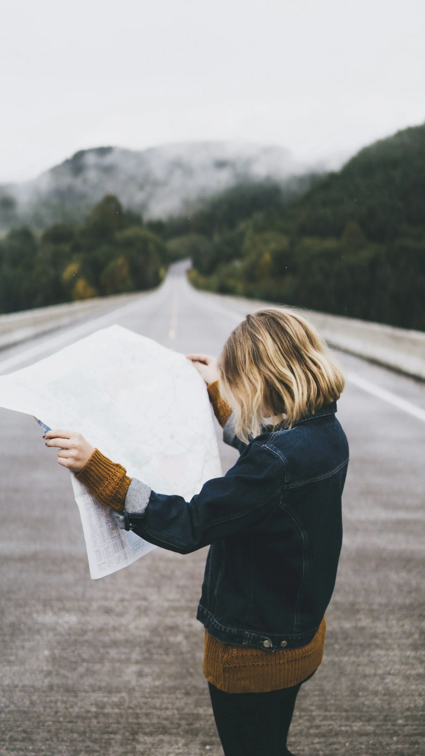 A woman with blonde hair holding a map and a pen standing on an empty road in a mountainous area with trees.