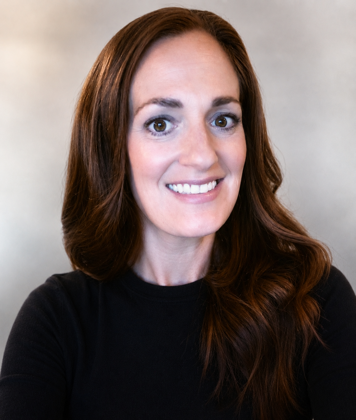 A woman with long, wavy red hair and brown eyes smiling at the camera, wearing a black top against a plain background.