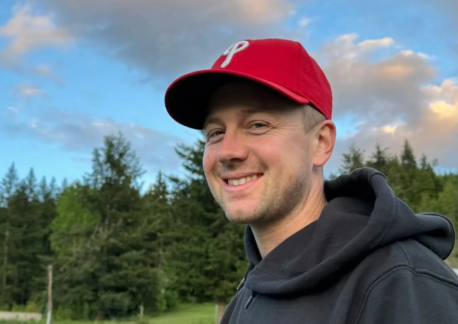 Sean Lowe, Owner/Operator, smiling wearing a red baseball cap and a black hoodie, outdoors with green trees and a partly cloudy sky in the background.