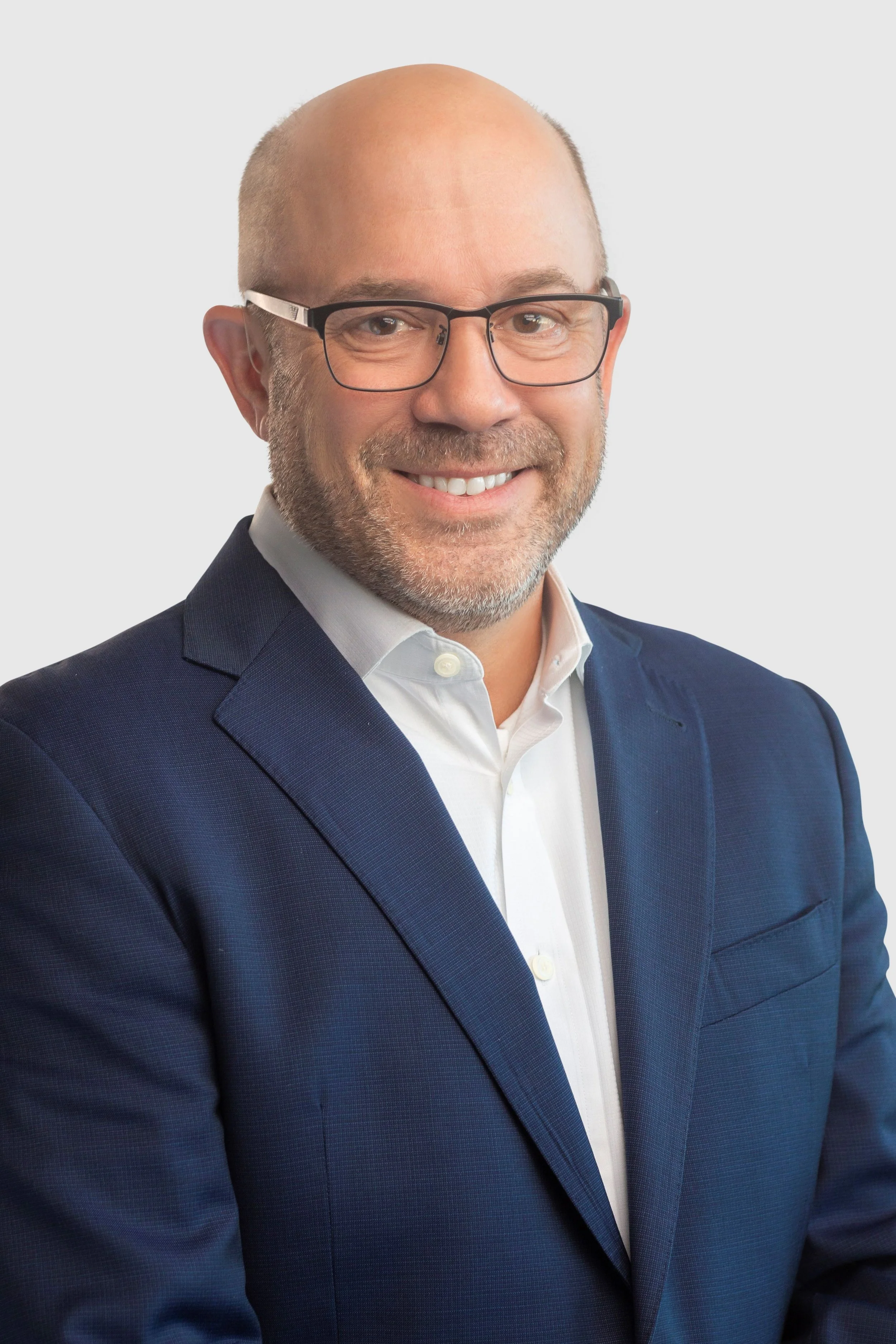 Headshot of a man with glasses, a bald head, and a gray beard, wearing a navy suit and white shirt, smiling against a plain background.