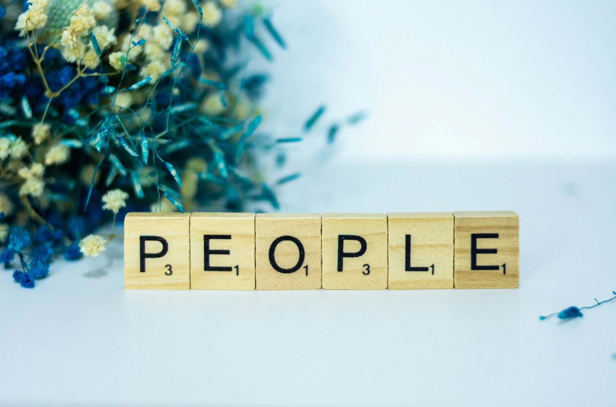 Close-up of wooden Scrabble tiles spelling the word 'PEOPLE' on a white surface, with yellow and blue flowers blurred in the background.