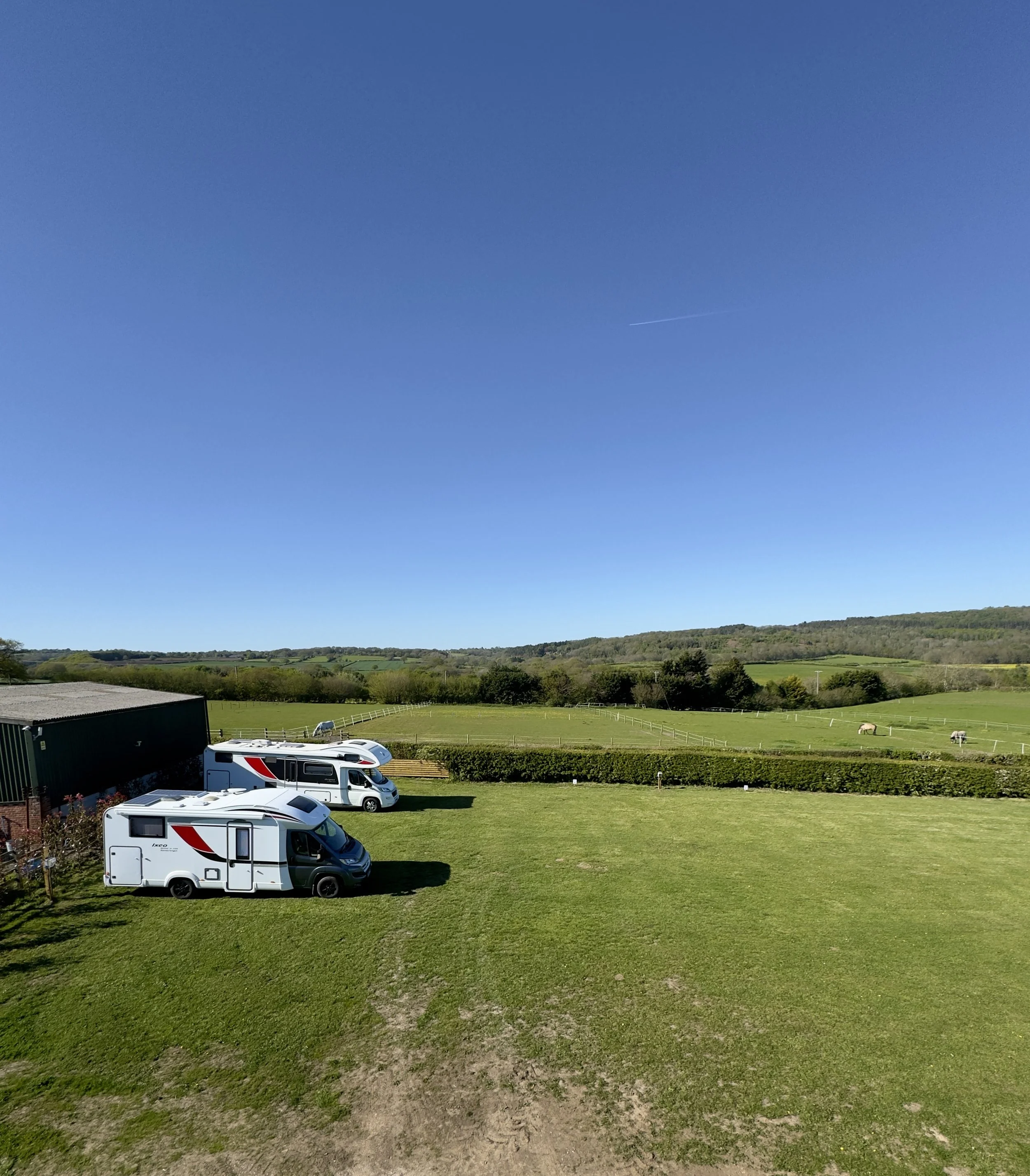 Green grassy landscape with three parked recreational vehicles near a building on the left, with rolling hills and trees in the background under a clear blue sky with a visible contrail.