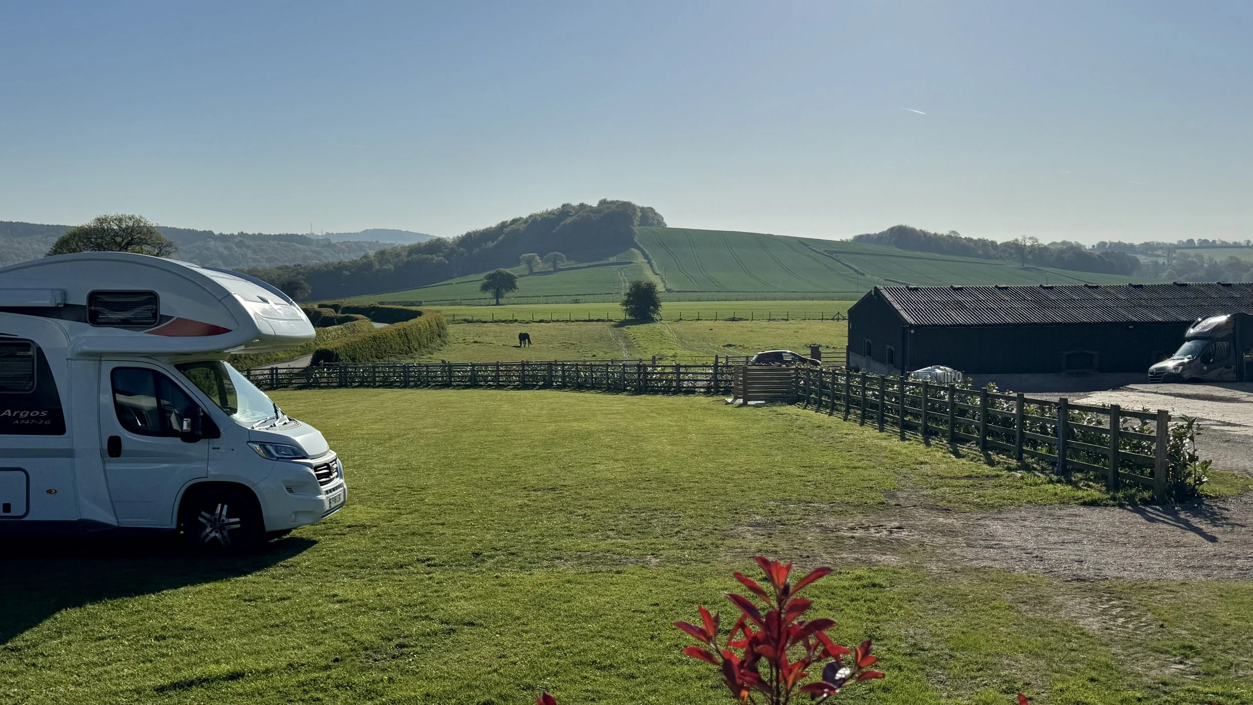 A scenic countryside with rolling green hills, a few trees, a horse grazing, and a small building. Two camper vans are parked on a grassy area in the foreground.