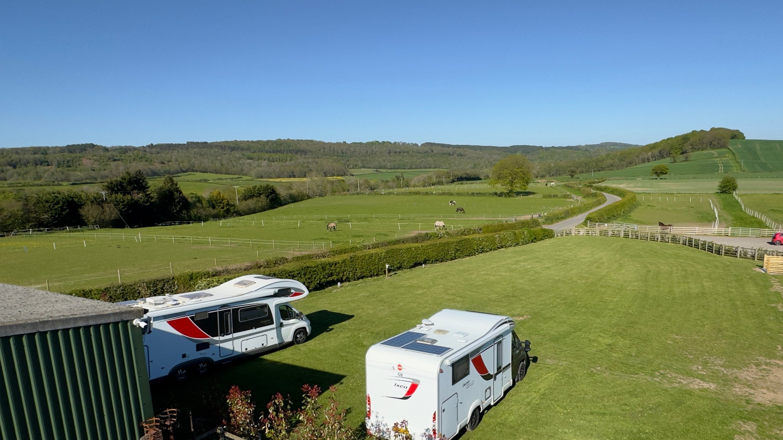 A scenic view of a rural landscape with green fields, trees, and rolling hills under a clear blue sky. Several motorhomes or campervans are parked on the lawn in the foreground.