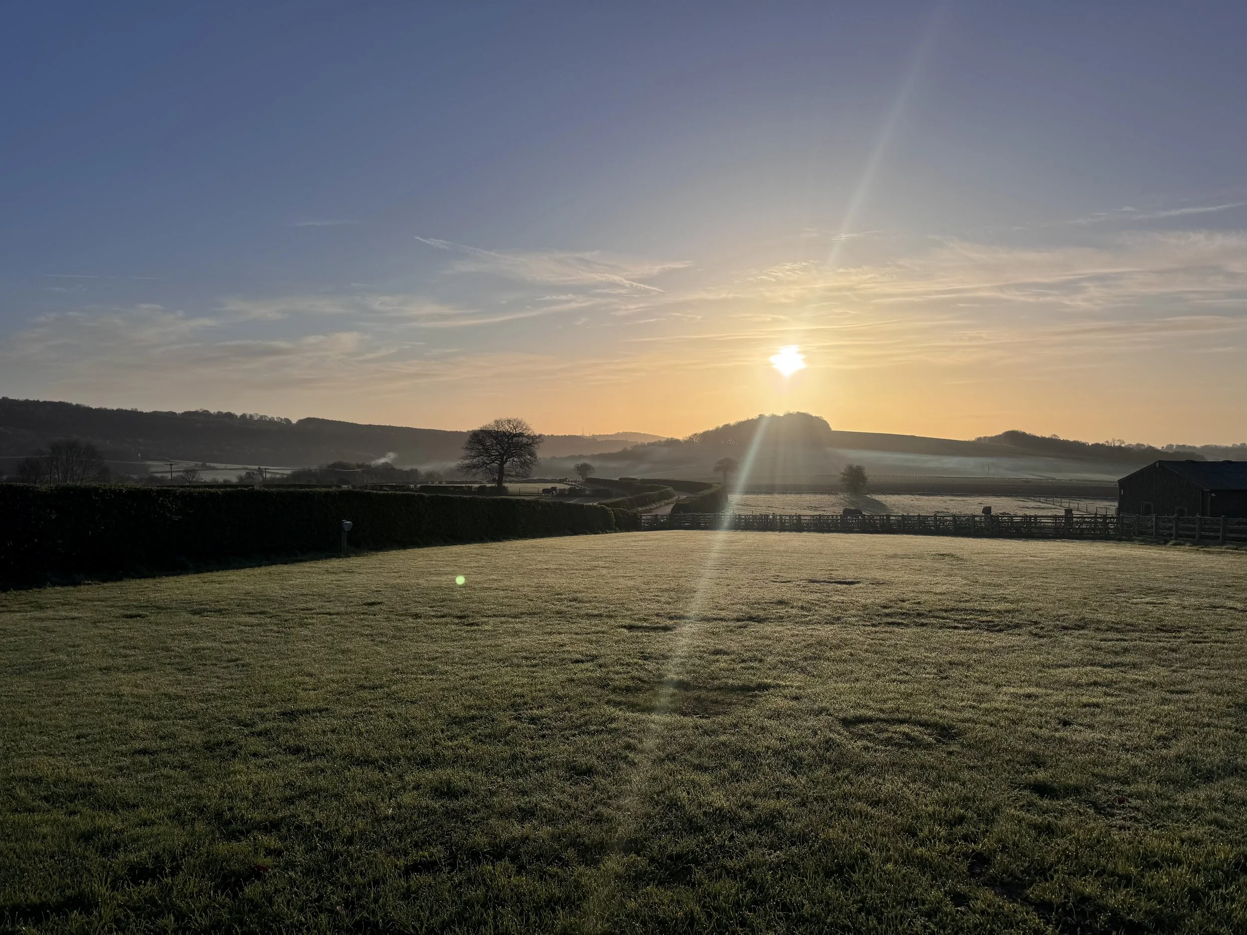 Sunrise over a rural landscape with rolling hills, trees, a farm building, and a fenced pasture.