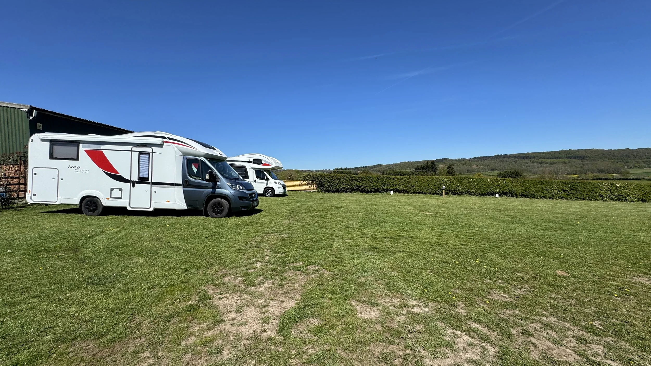 two motorhomes parked on a grassy field near a green shed, with rolling hills and a clear blue sky in the background.
