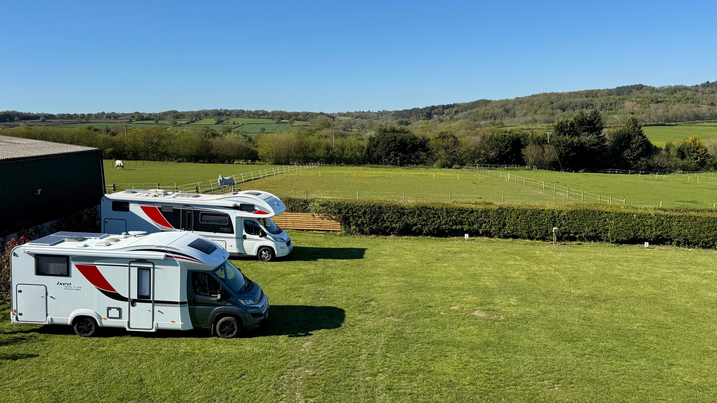 Three white motorhomes parked on a grassy field with a hedge in the foreground, and rolling green hills, trees, and a clear blue sky in the background.