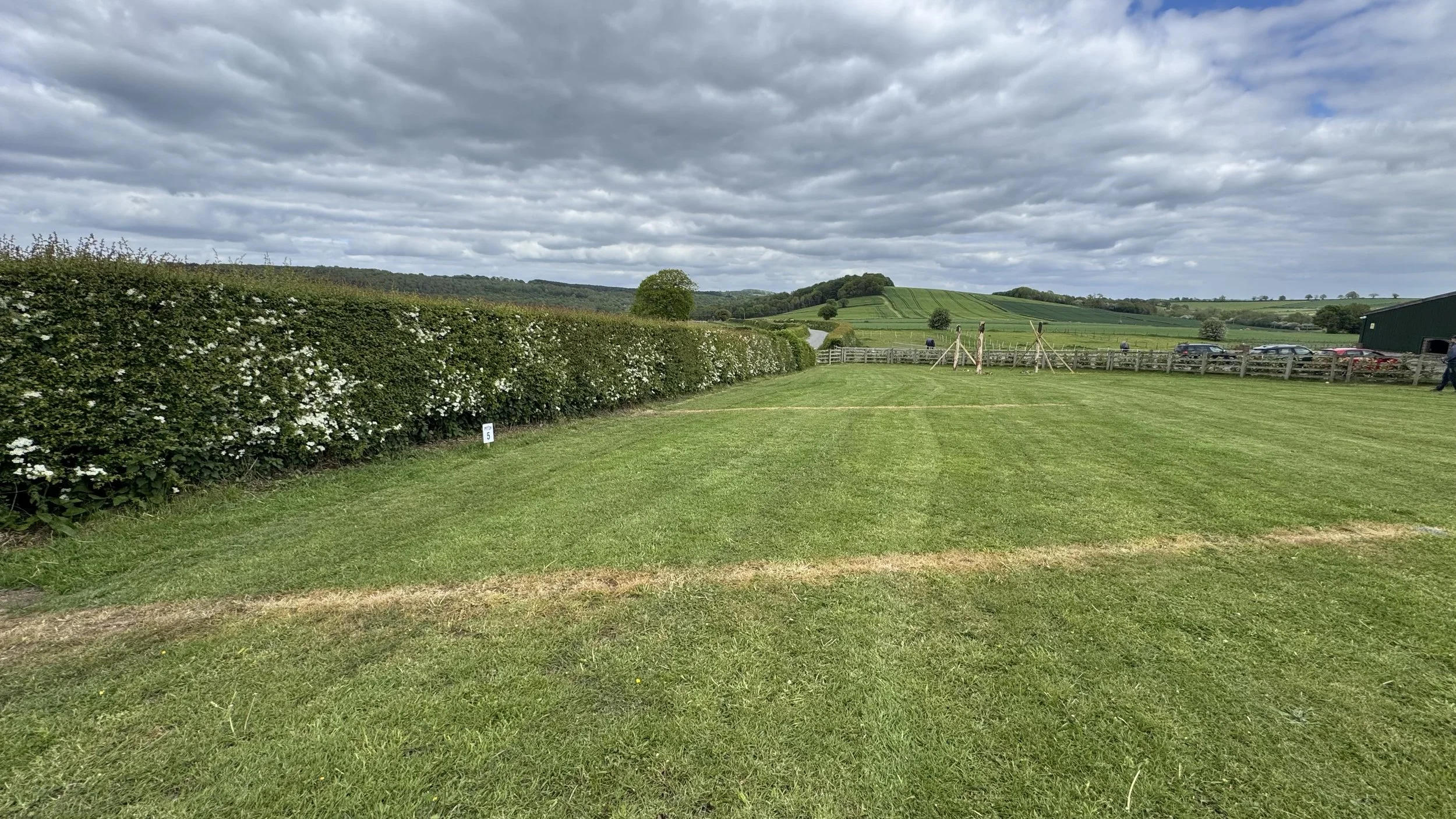 A green open field with a hedge on the left, trees, rolling hills, and a cloudy sky in the background.