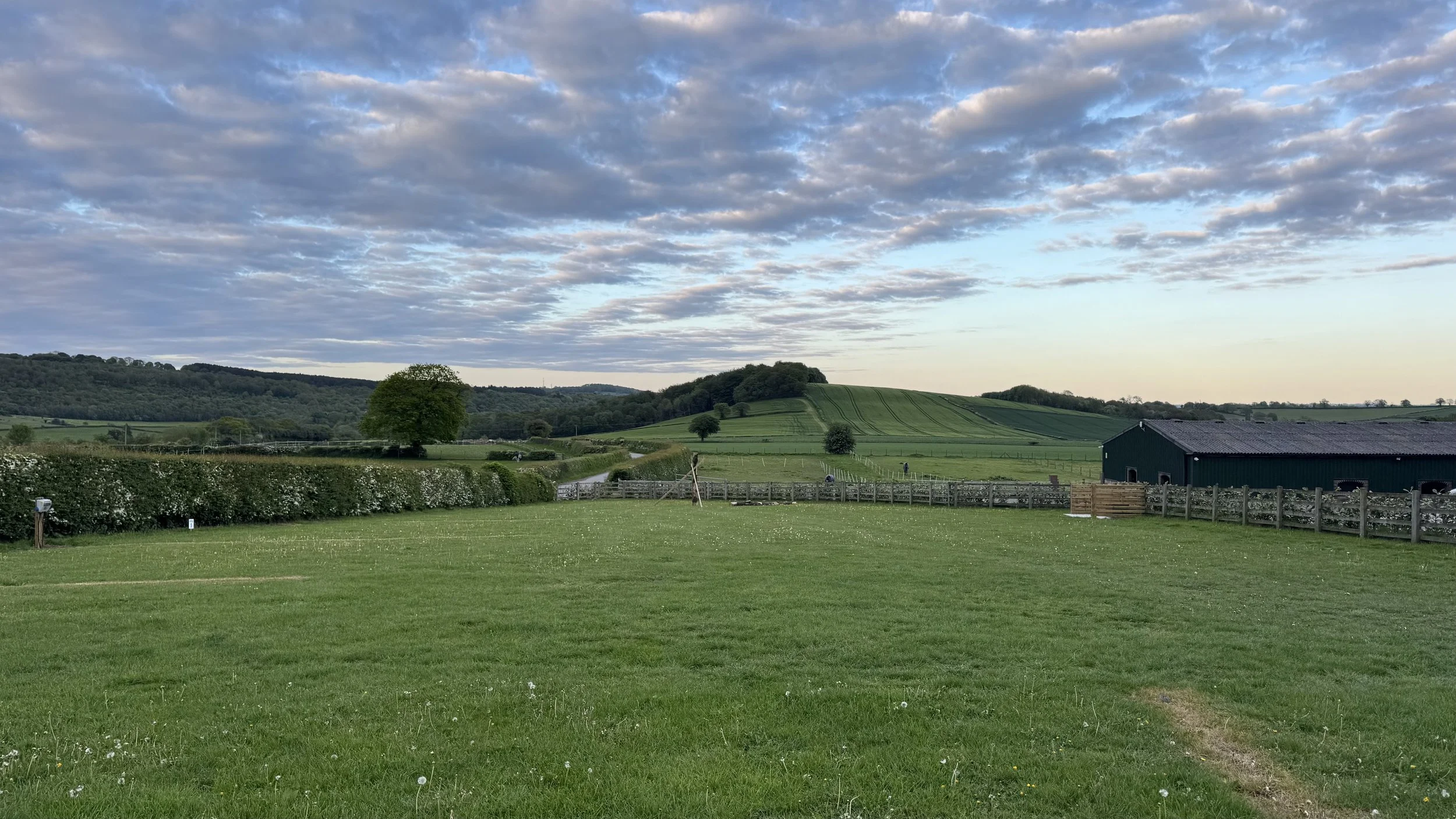 A scenic view of a lush green rural landscape with rolling hills, trees, a wooden fence, a barn, and a sky filled with scattered clouds in the distance.
