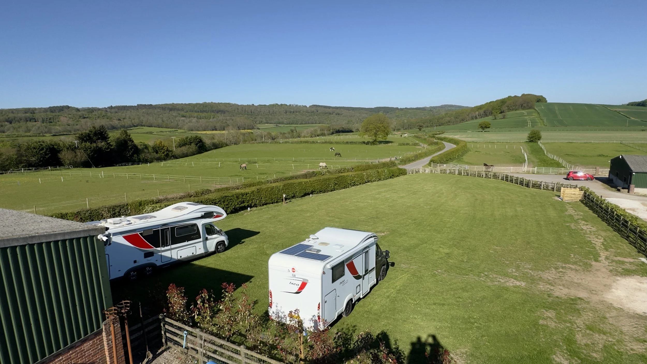A scenic countryside view with green fields, rolling hills, and a clear blue sky. Several caravans and motorhomes are parked near a house, and a few horses graze in the fenced fields.
