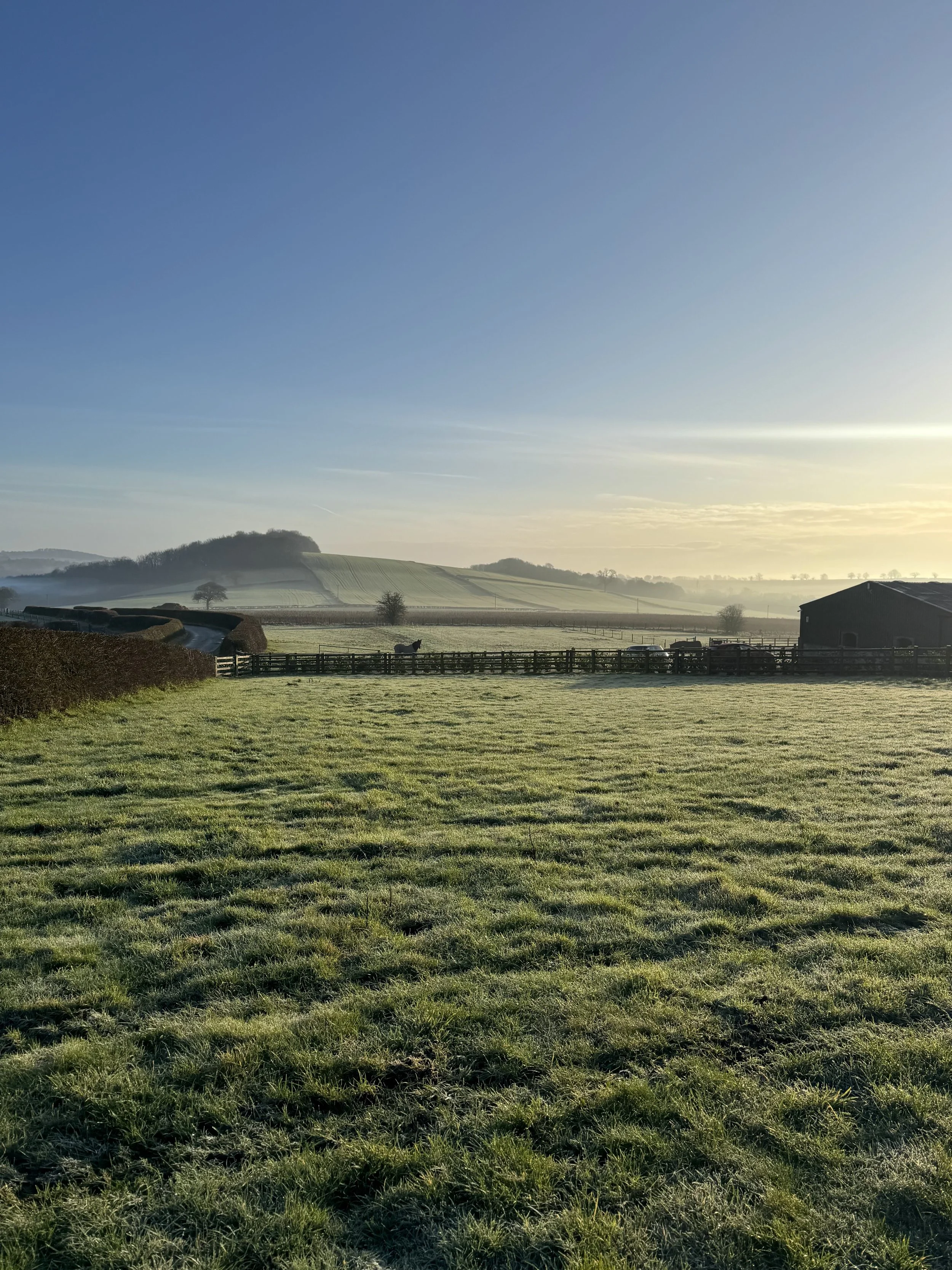 A peaceful countryside landscape during sunrise, featuring green fields, a wooden fence, distant trees, and rolling hills under a clear blue sky with some clouds.