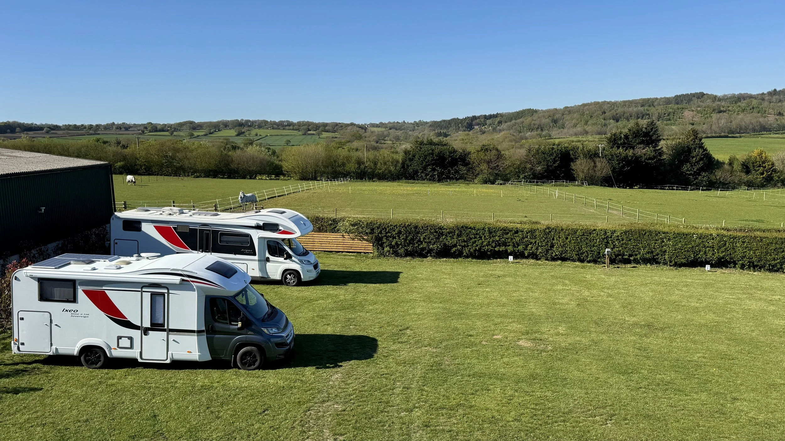 Two motorhomes parked on a grassy area with a black building on the left, set against a backdrop of green fields, trees, and rolling hills under a clear blue sky.