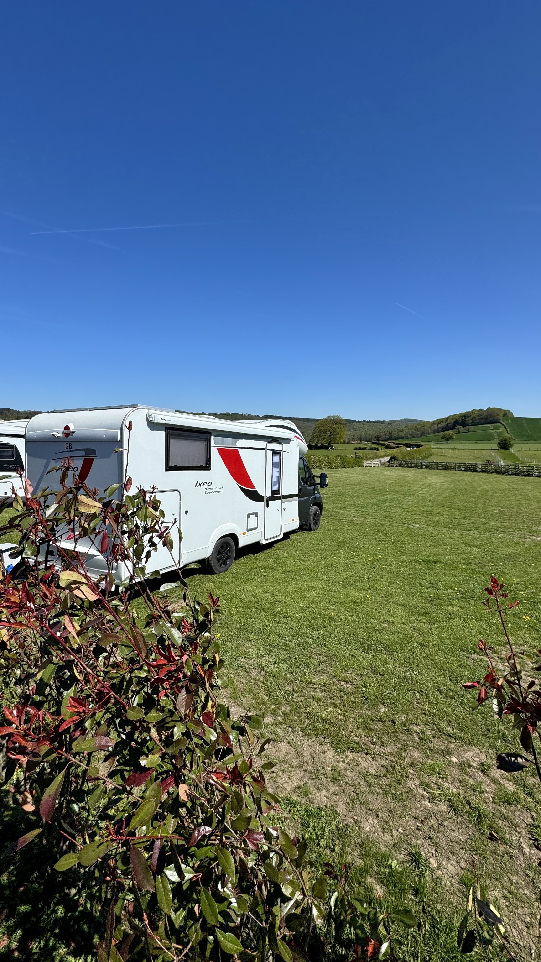 A white motorhome parked on a grassy field under a clear blue sky, with rolling green hills in the background and some red and green foliage in the foreground.