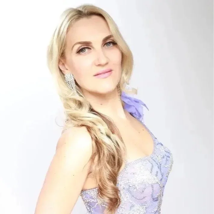 A woman with long blonde hair styled in loose waves, wearing a lavender lace dress with a purple ribbon on her shoulder, and large sparkling earrings, posing against a white background.