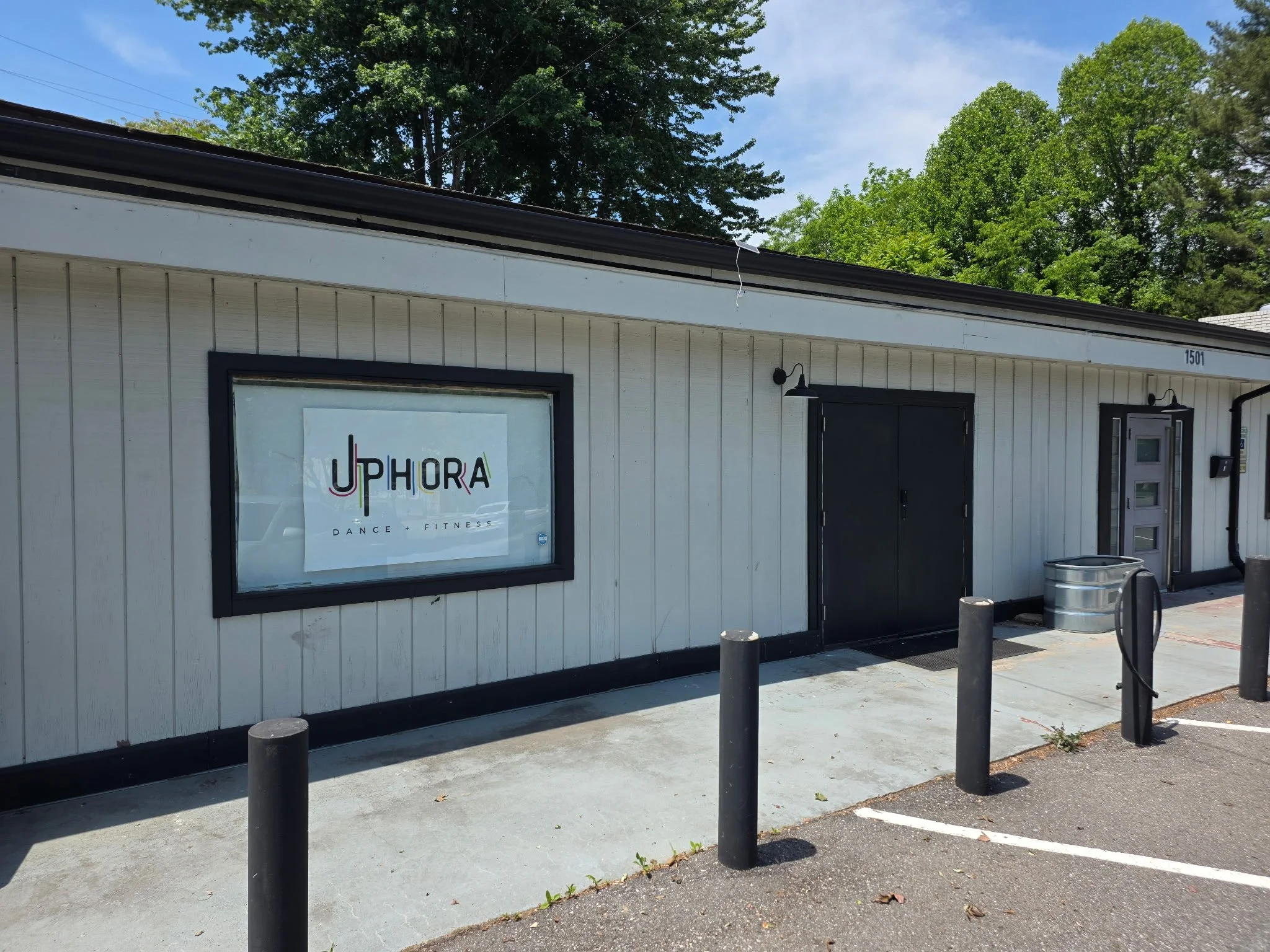 Exterior of a building with a sign that reads 'Uphora Dance + Fitness', black door, and outdoor lighting fixtures. Several black bollards and trash cans are in front of the building. Trees with green foliage are in the background under a blue sky.
