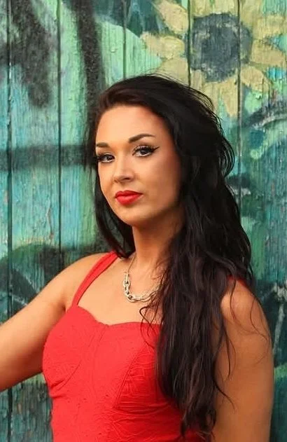 A woman with long dark hair and red lipstick posing in front of a colorful, painted wooden fence.
