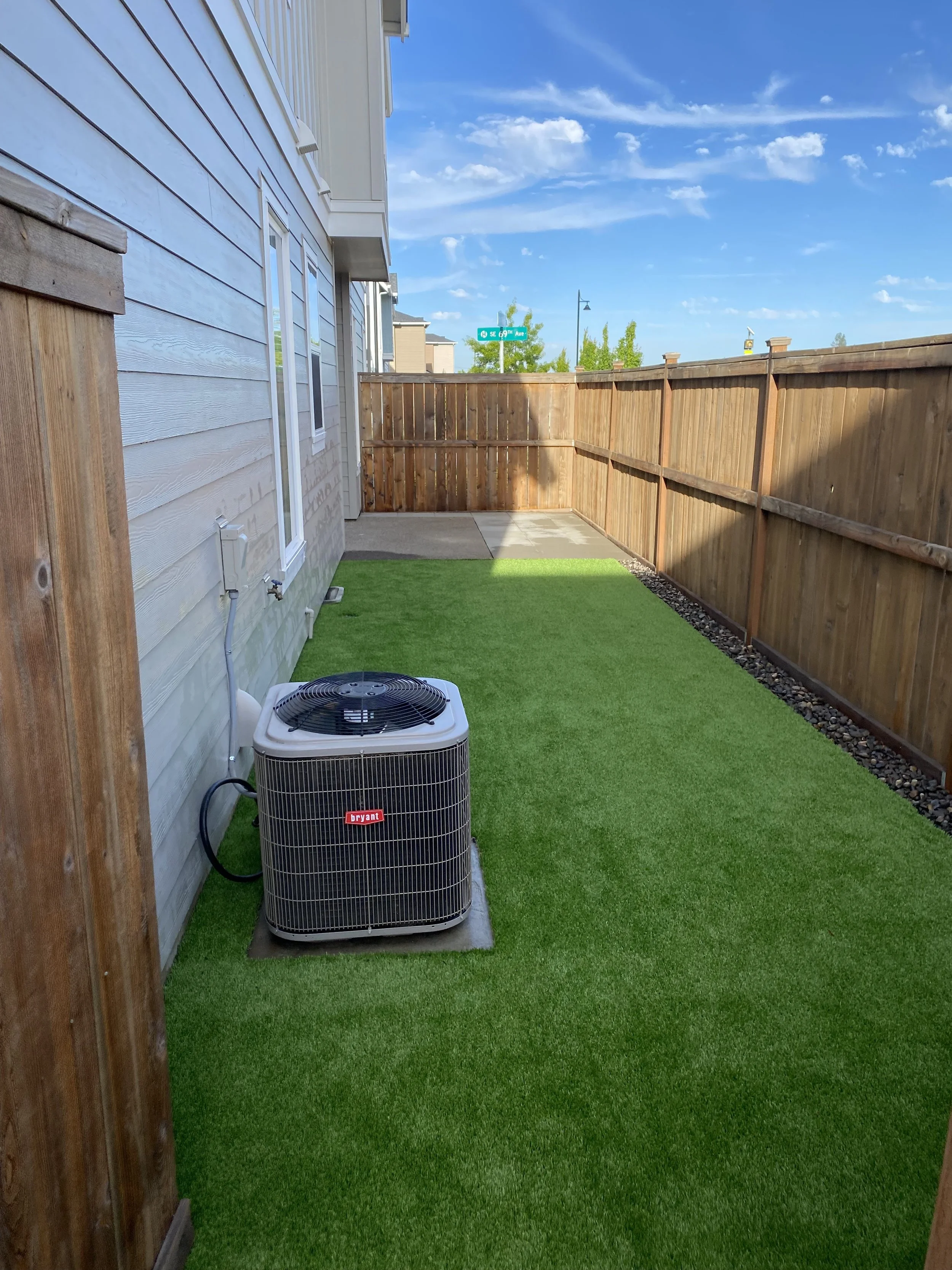 Small backyard with artificial grass, a wooden fence, and a Bryant air conditioning unit near a house with gray siding and white trim. Clear blue sky with a few clouds.