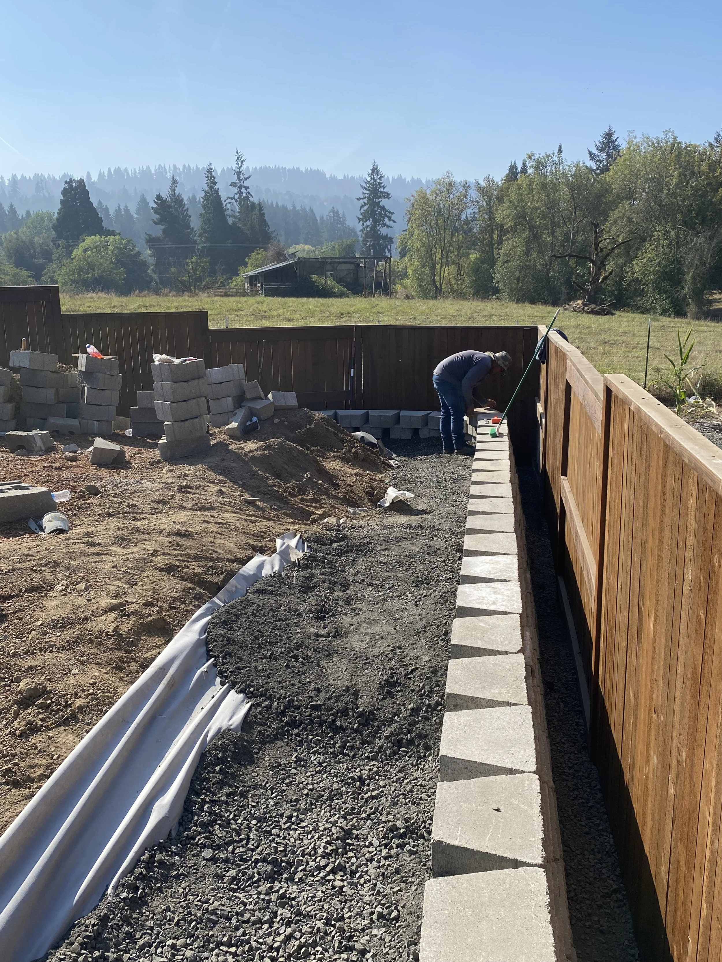 A construction worker placing concrete pavers along a wooden fence in a backyard under cloudy sky with trees in the background.