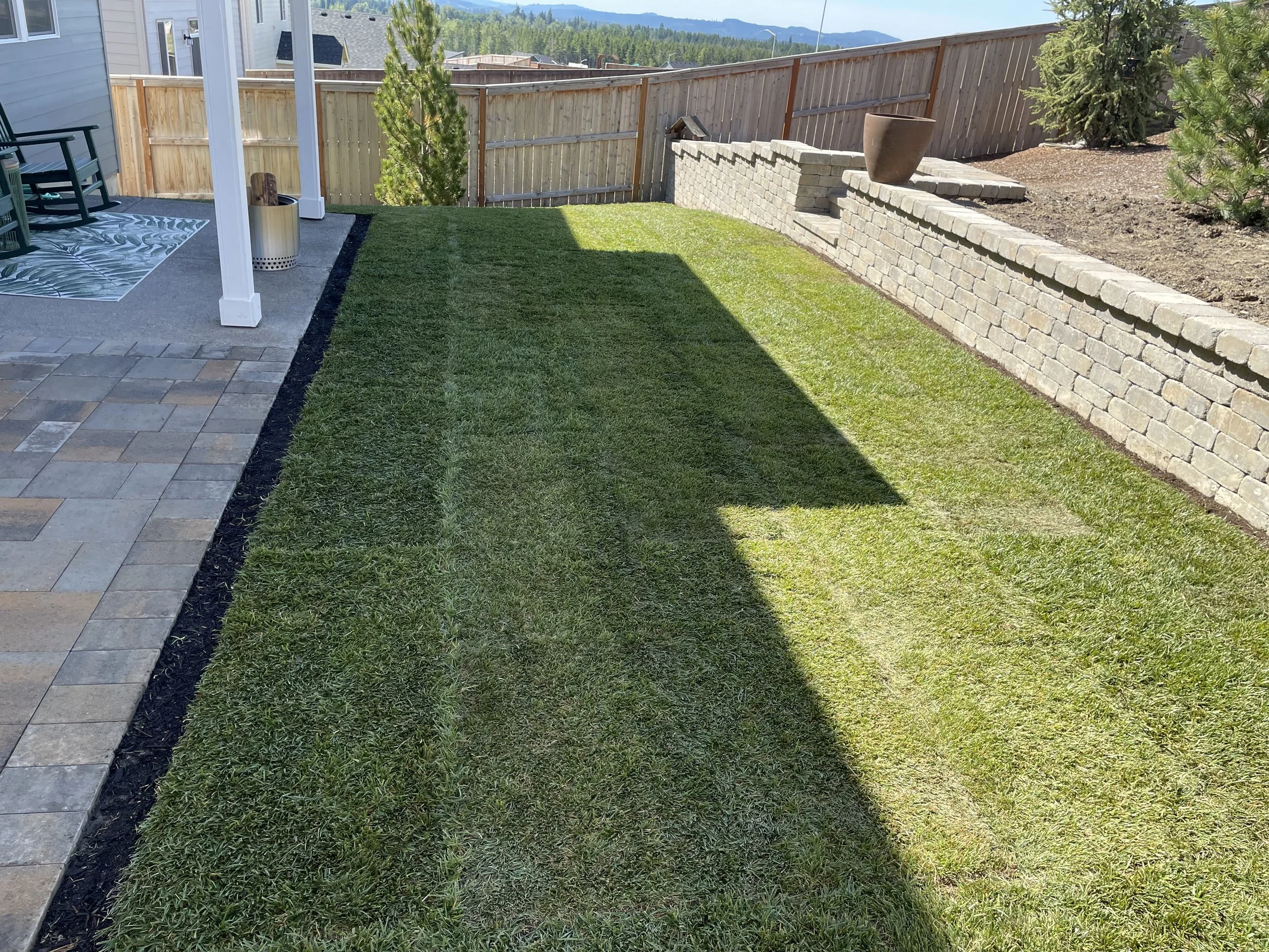 A backyard with a small patch of grass bordered by a concrete patio, a wooden fence, and a retaining wall with potted plants. In the background, there are mountains and clear skies.