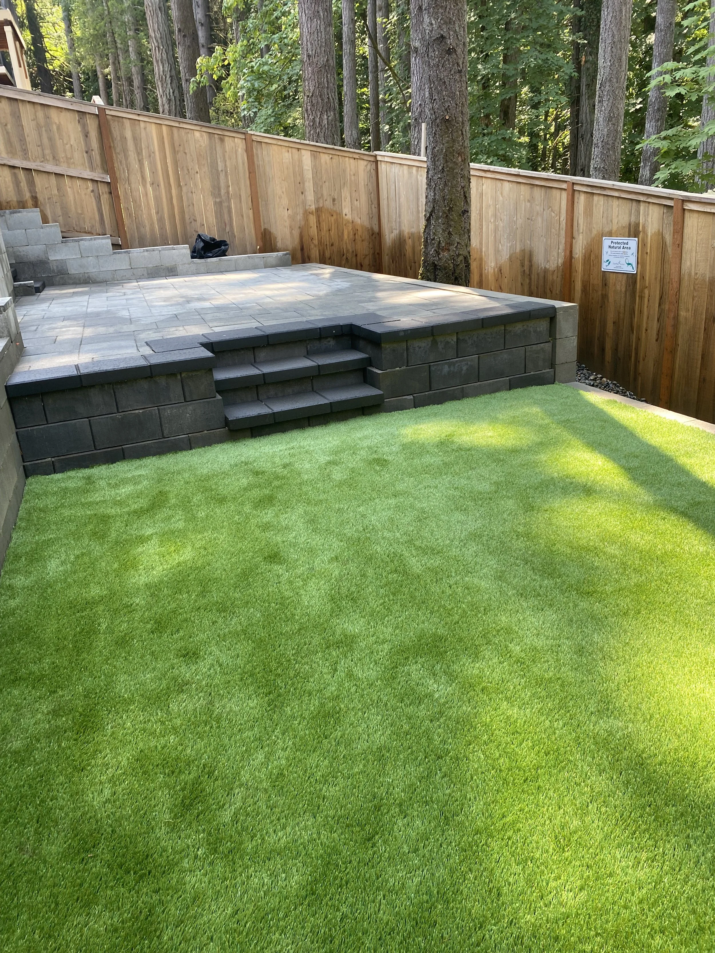 A backyard with green artificial turf, a raised patio with gray pavers, a wooden privacy fence, and surrounded by forest trees.