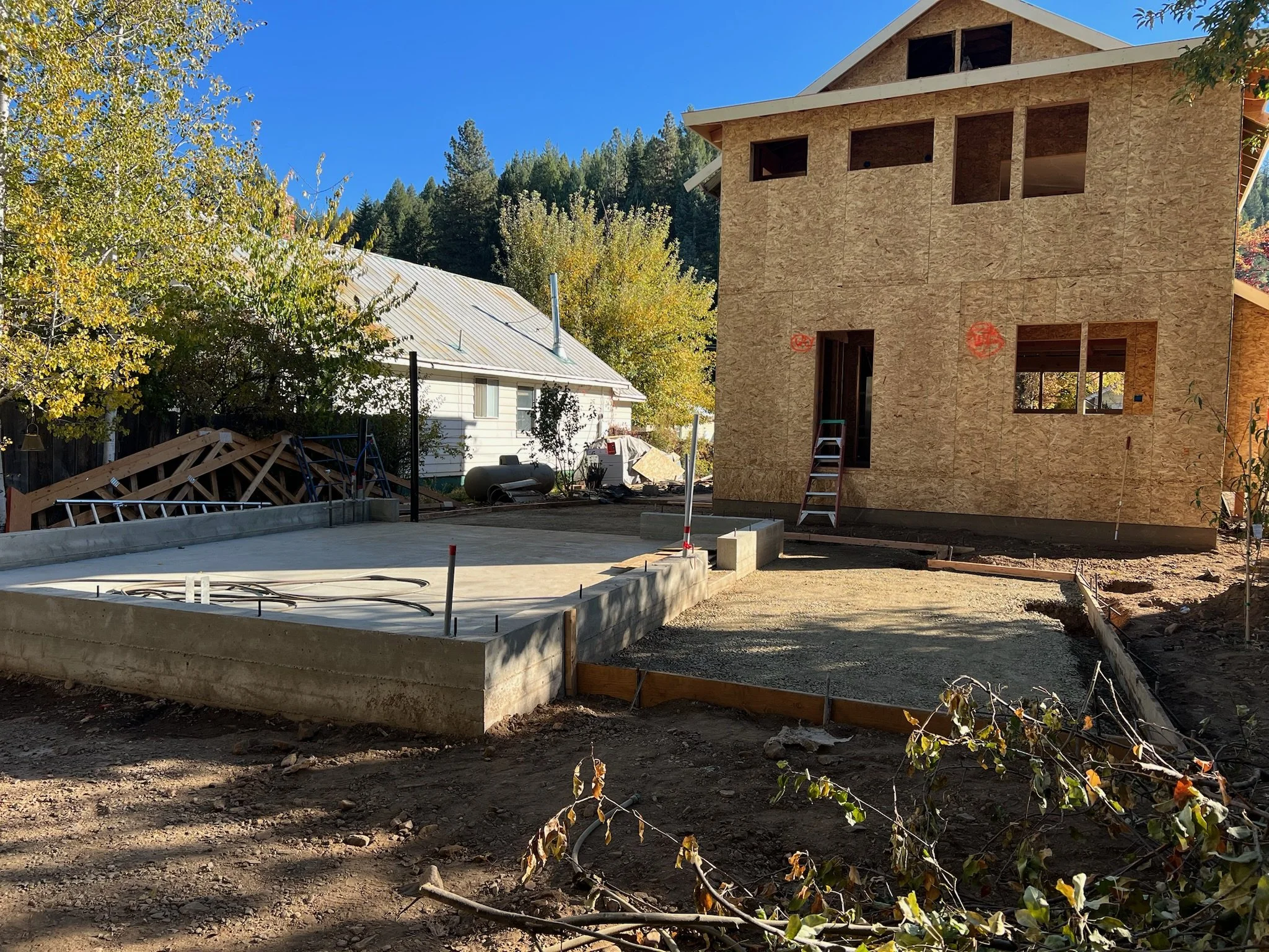 Construction site with a two-story house under construction, plywood walls, ladder, and concrete foundation, surrounded by trees and a clear blue sky.