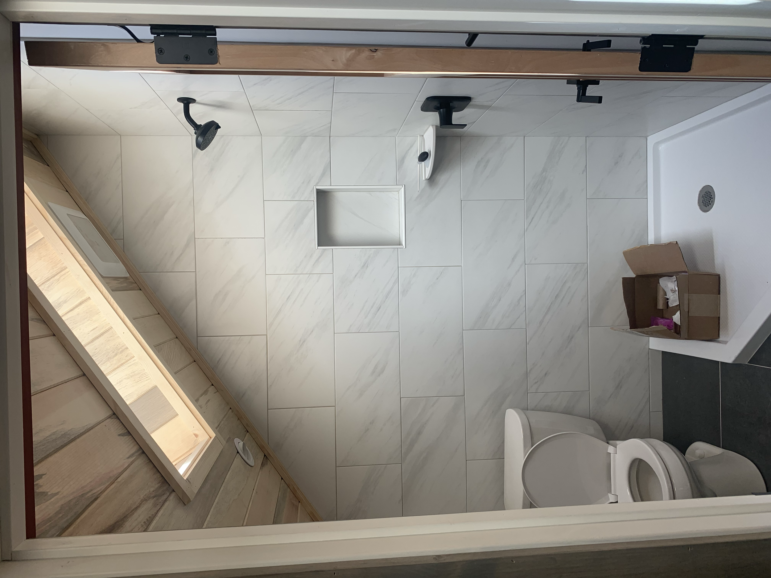 View of a bathroom with white marble tiles, a shower head, a soap dish, a white toilet, and a cardboard box on the floor.