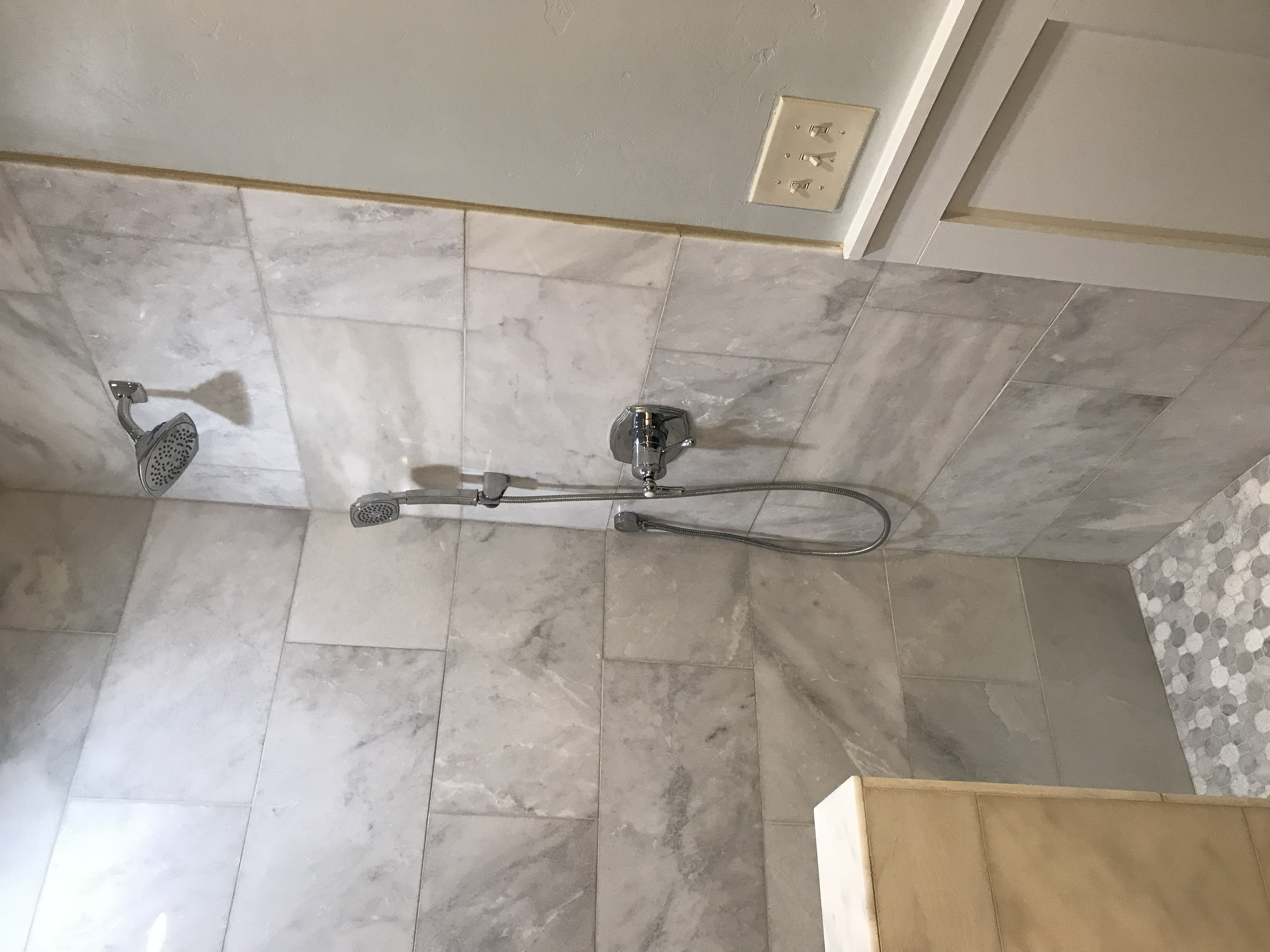 Bathroom shower with two showerheads, tiled wall, electrical outlet, and a bench, with mosaic tiles on the floor.