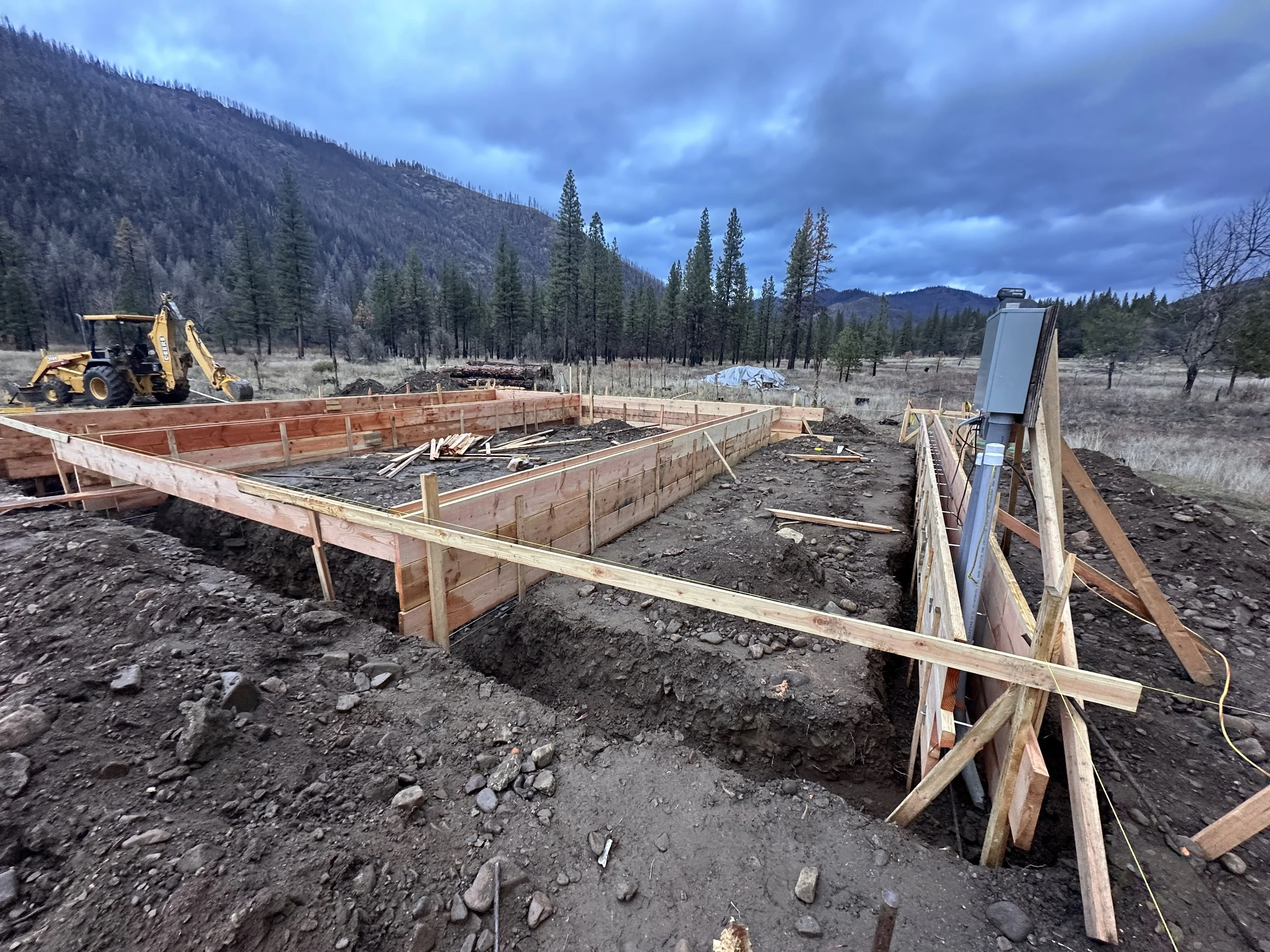 Construction site with wooden framing for a foundation, a backhoe, and electrical panel in a mountainous area with trees and cloudy sky.