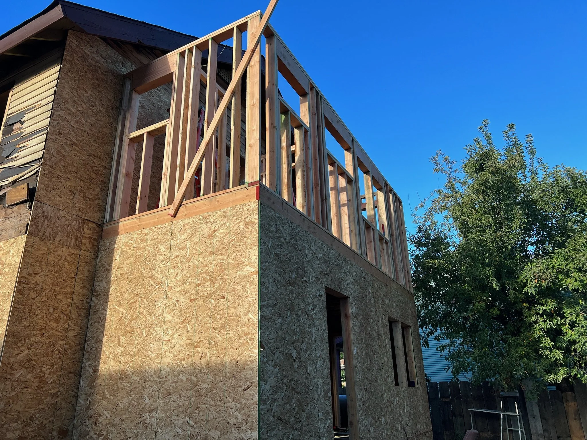 A house under construction showing wooden framing and exterior wall sheathing with plywood boards. The sky is clear and blue, and a tree is visible on the right side.