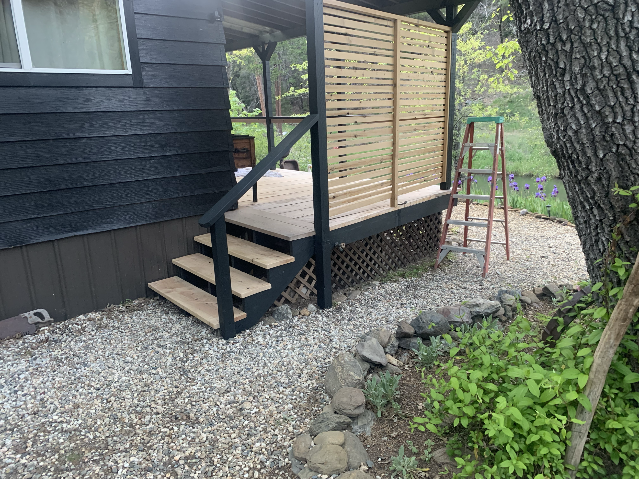 A small deck at an outdoor area with a black exterior wall and a beige wooden privacy screen. A red and silver ladder leans against the deck, with large trees and purple flowers visible nearby.