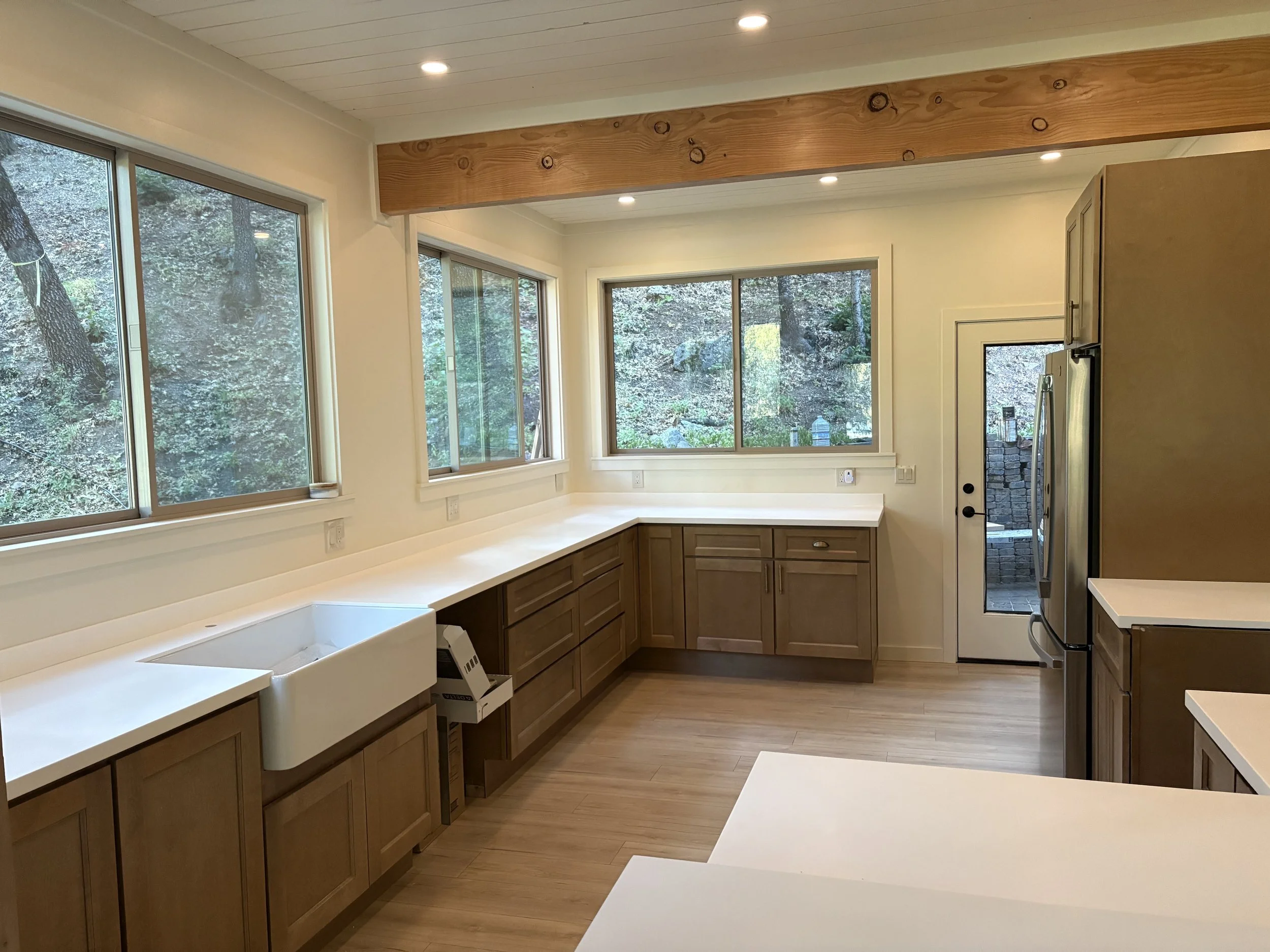 A kitchen with large windows showing trees outside, wooden cabinets, a white countertop, and a white farmhouse sink.