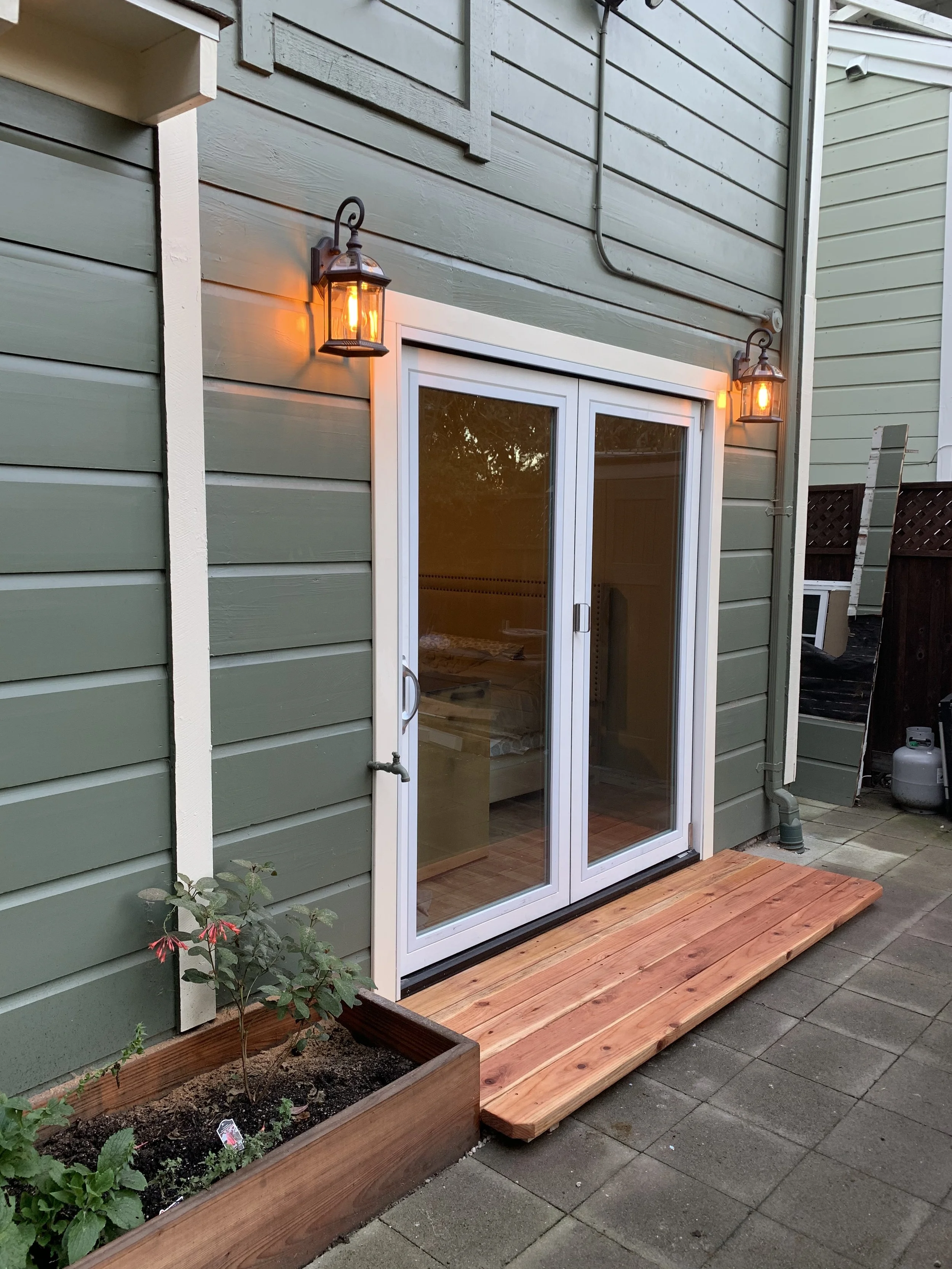 Exterior view of a green house with sliding glass door, wooden deck, and two outdoor wall-mounted lantern lights that are turned on. There is a flower bed with plants in front of the house.