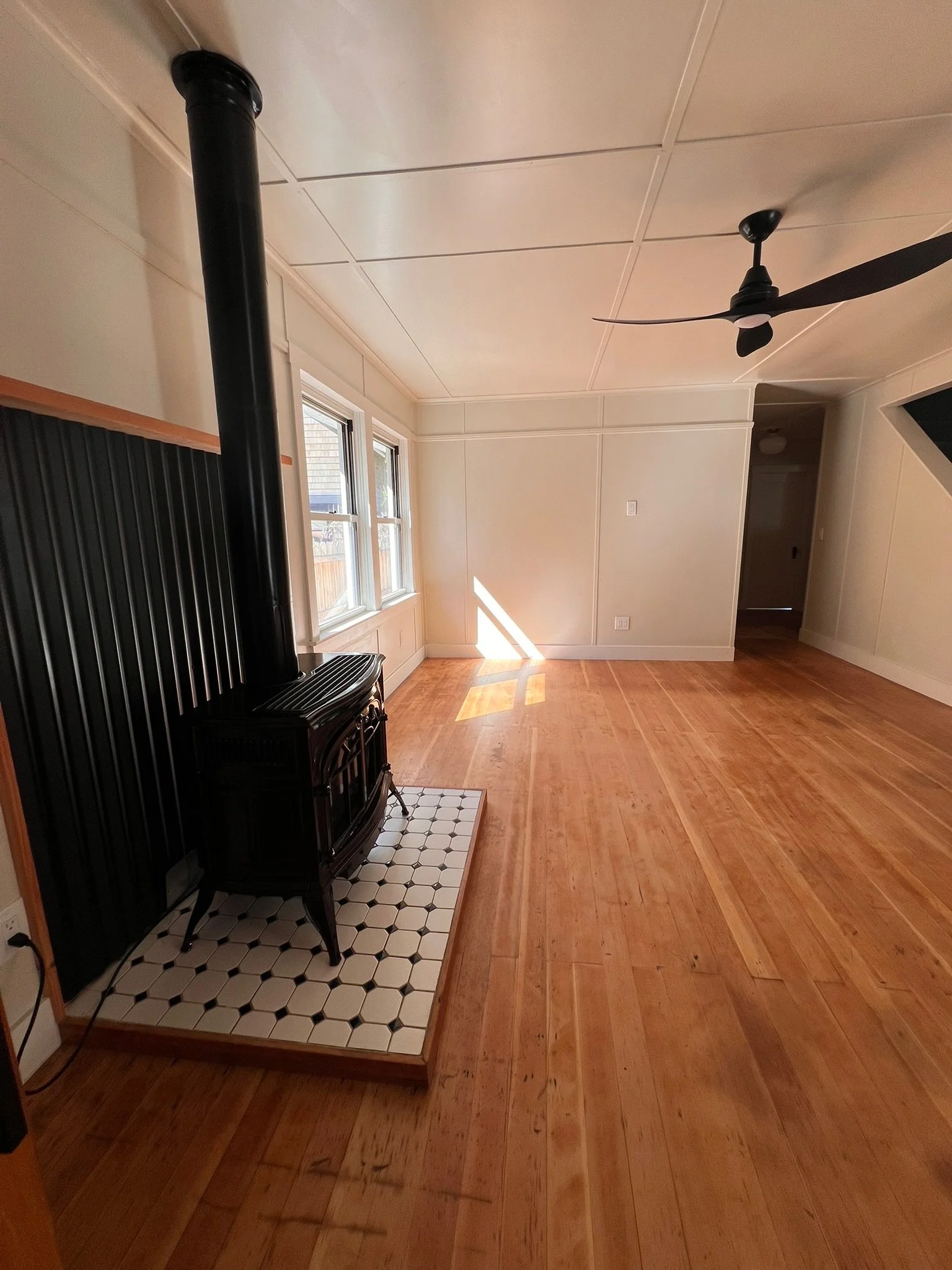A room with wooden floors, a black wood-burning stove on a tiled hearth, two windows letting in natural light, a ceiling fan, and a doorway leading to another room.
