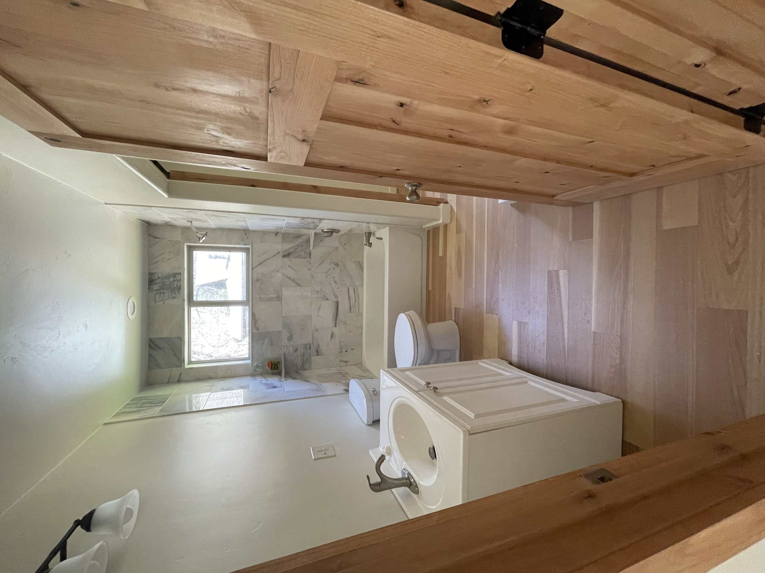 A view of a bathroom with a wooden door, a window, a bathtub, and a toilet, with light-colored walls and wood flooring