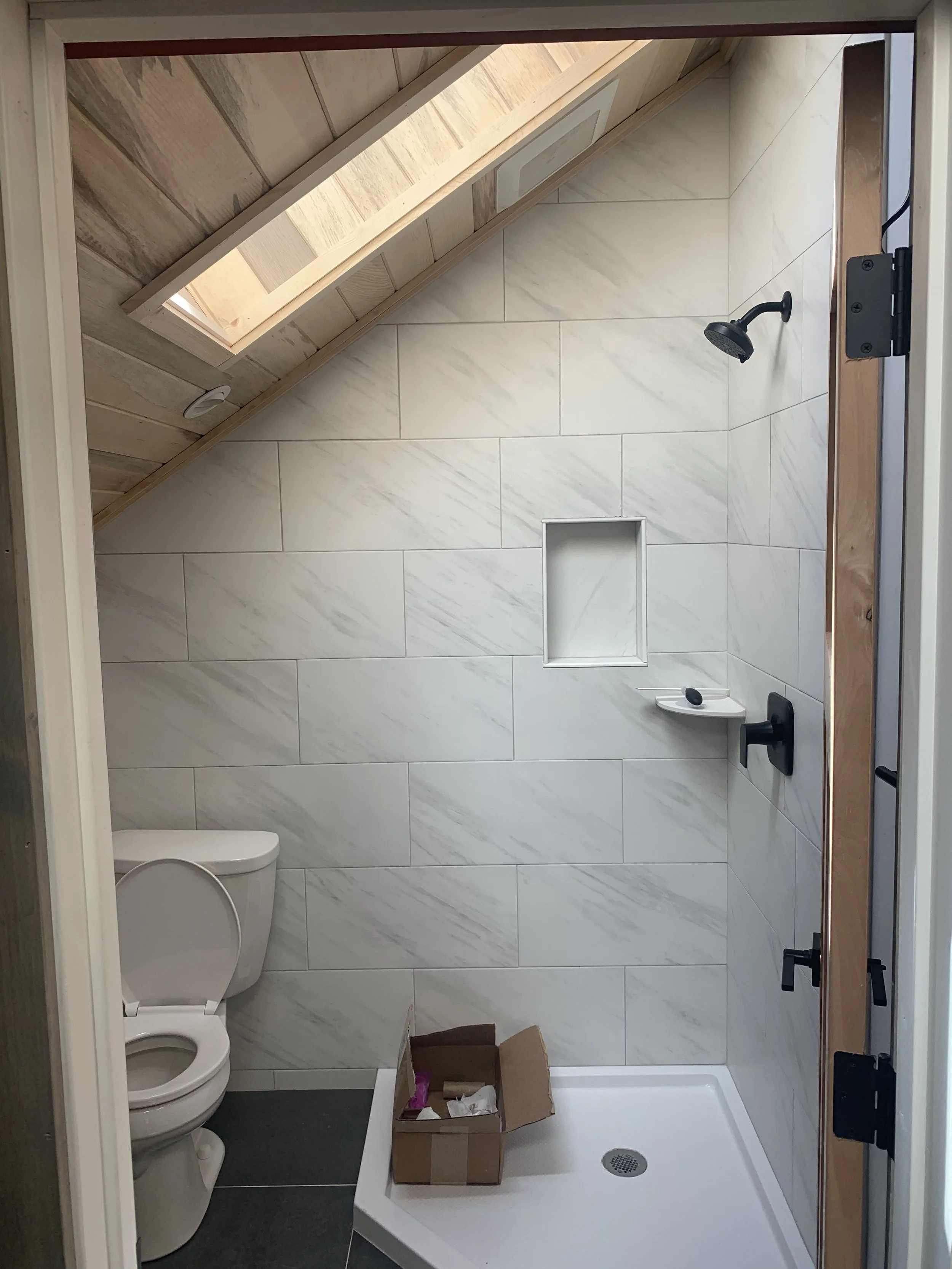 Bathroom with white marble tiles, a toilet, a shower with a black showerhead, a built-in shelf, roof window, and a cardboard box with items inside.