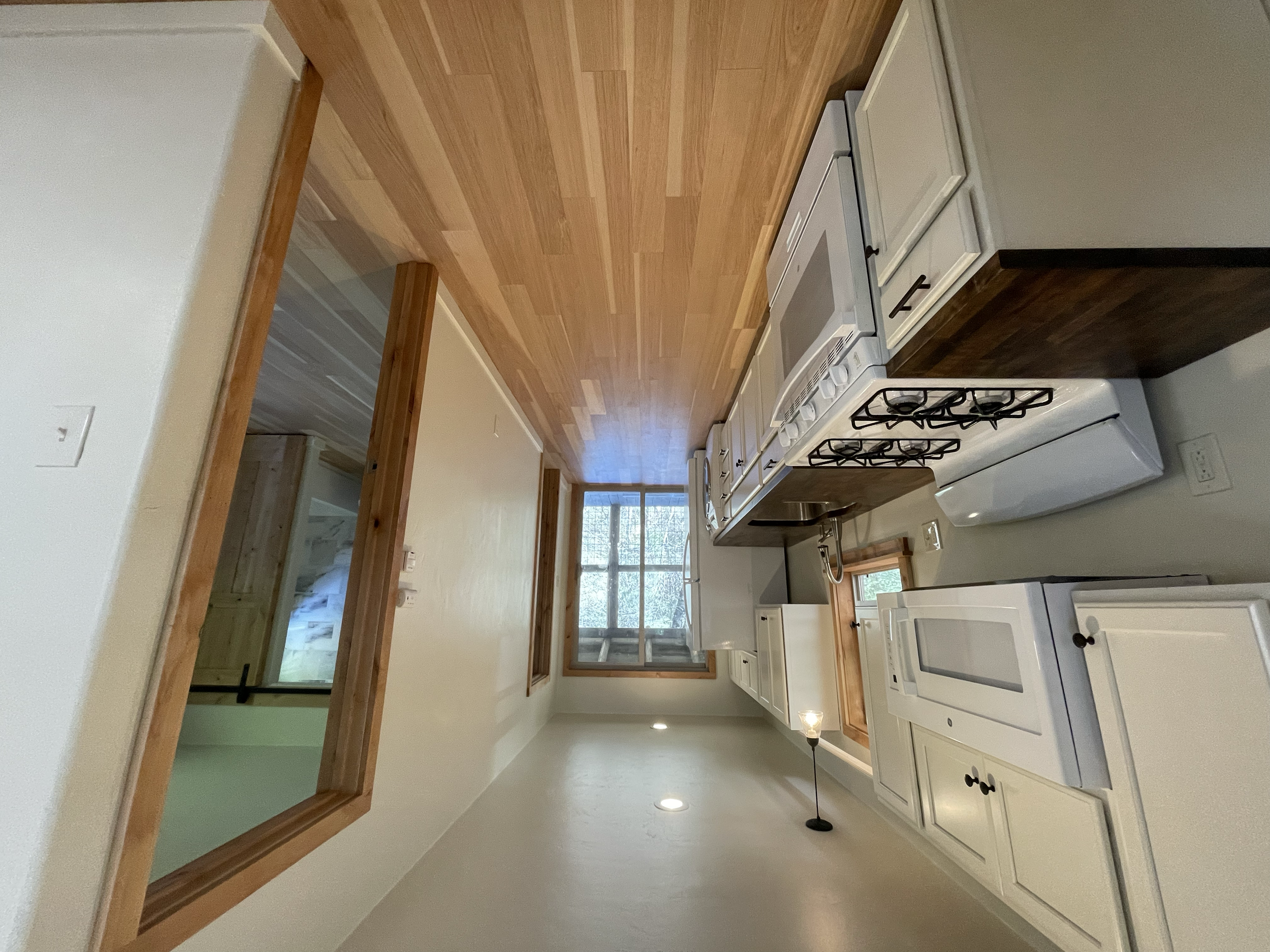 A kitchen with white cabinets, a stove, and a window overlooking the outdoors. The kitchen has light wood countertops and a wooden trim around a pass-through window.