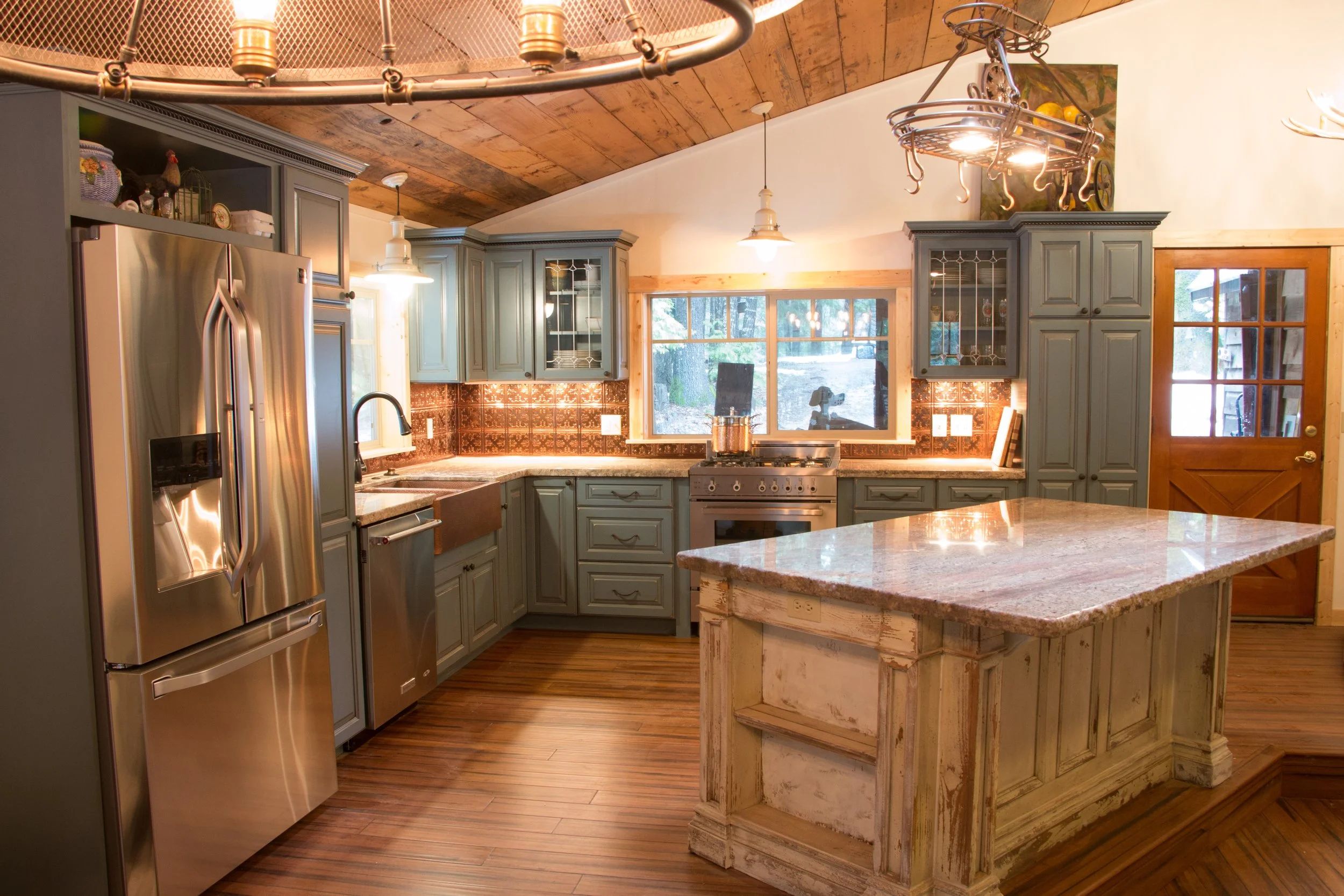 A kitchen with gray cabinets, a wooden ceiling, stainless steel refrigerator, granite countertop, a kitchen island with distressed wood finish, large window, pendant lights, and wooden door.