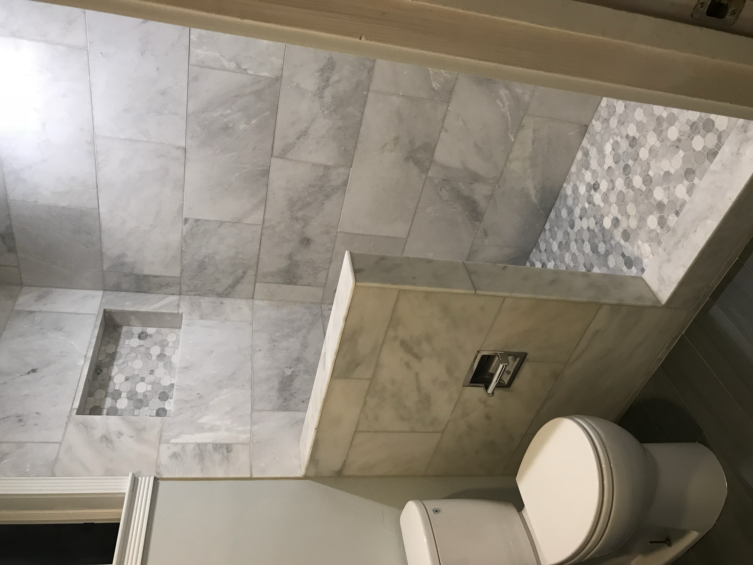 A bathroom shower area with grey marble tiles on the wall and hexagonal mosaic tiles on the floor of the shower. There are two recessed shelves in the shower wall. The lower corner of a white toilet is visible in the bottom right corner of the image.