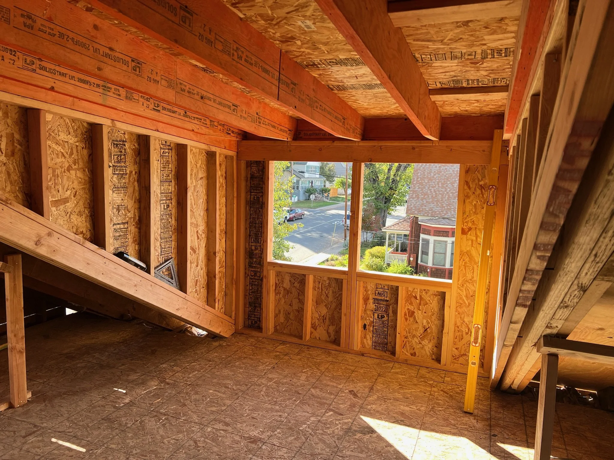 View of the interior of a house under construction, showing wooden framing and plywood sheathing, with a large window opening and a neighborhood visible outside.