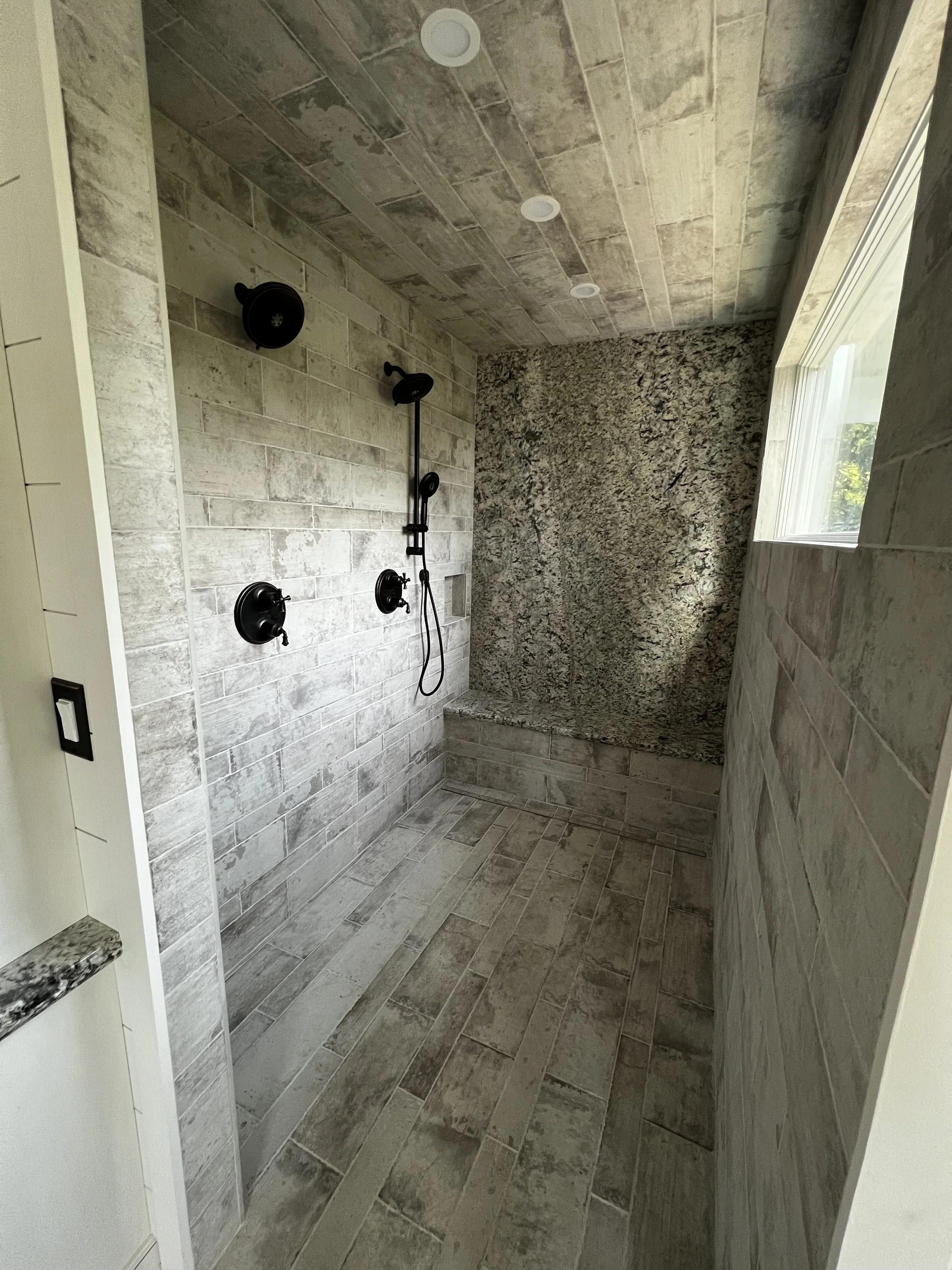 A spacious walk-in shower with gray stone tiles on the floor, walls, and ceiling. It features a black shower head, a handheld shower wand, and two black wall-mounted controls. A large window on the right side allows natural light to enter.