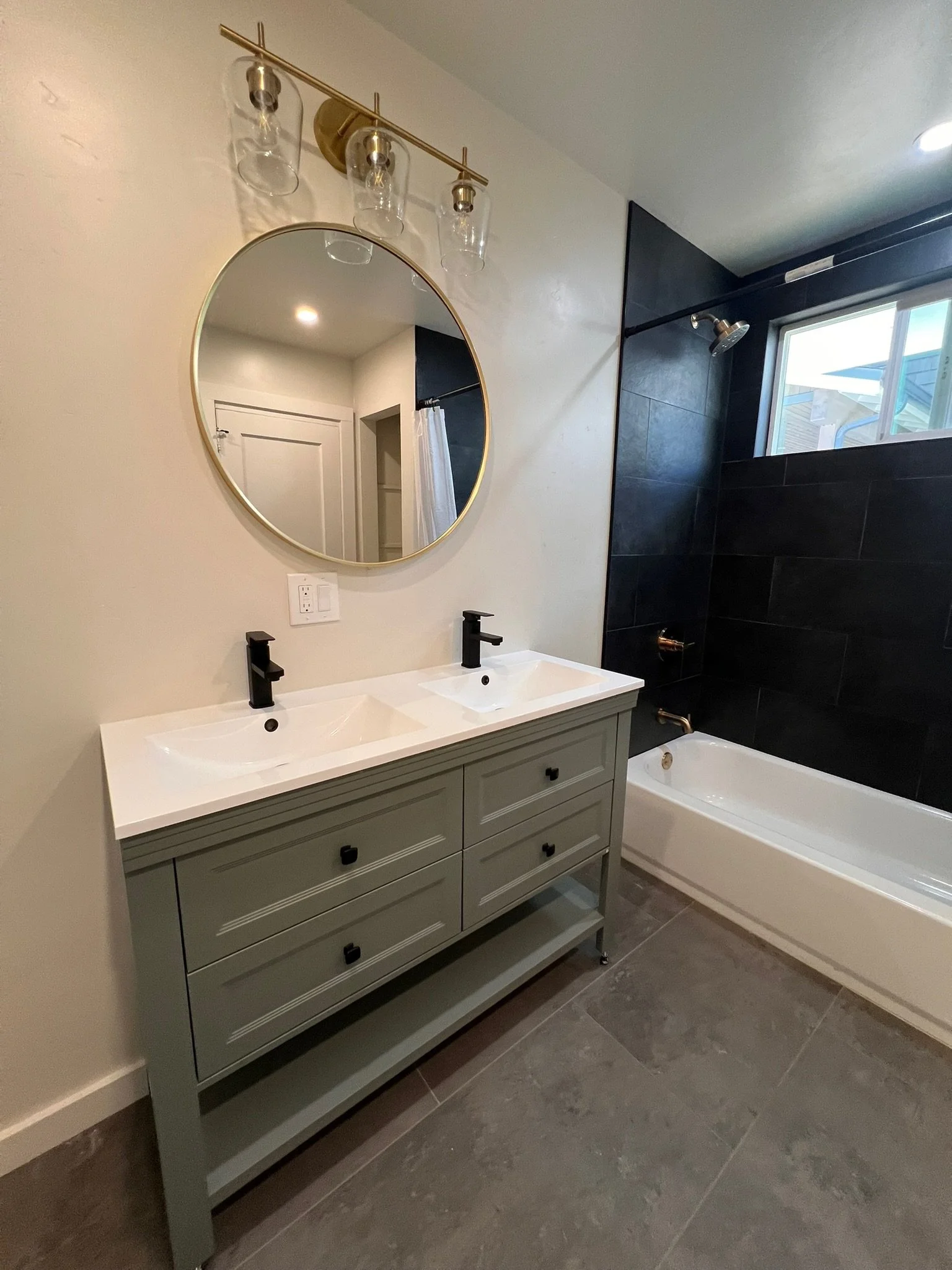 A bathroom with a double vanity, black faucets, a large round mirror, a white bathtub, and a black tiled shower area with a window.
