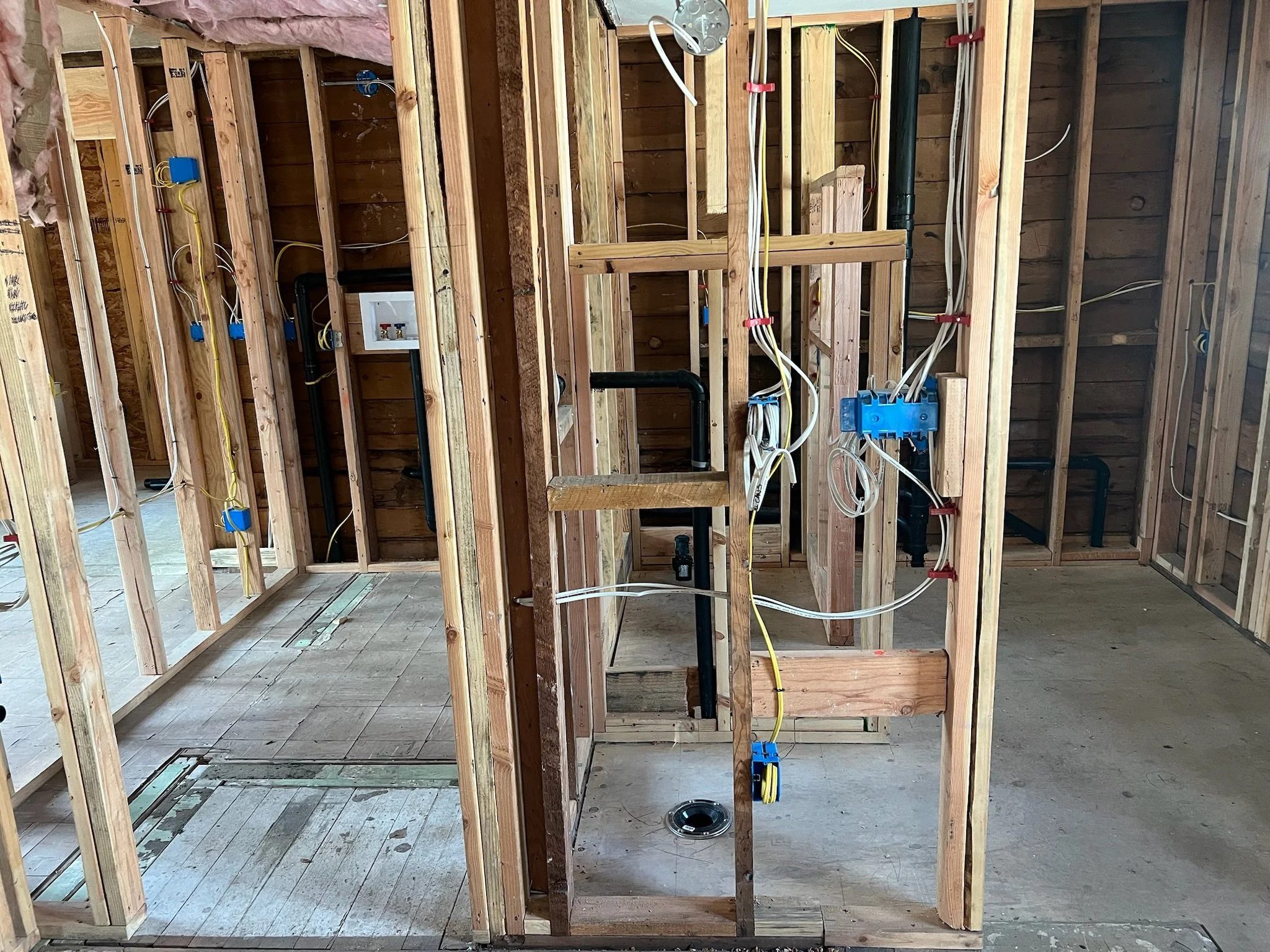 Interior framing of a house under construction, showing wooden studs, electrical wiring, and plumbing pipes.