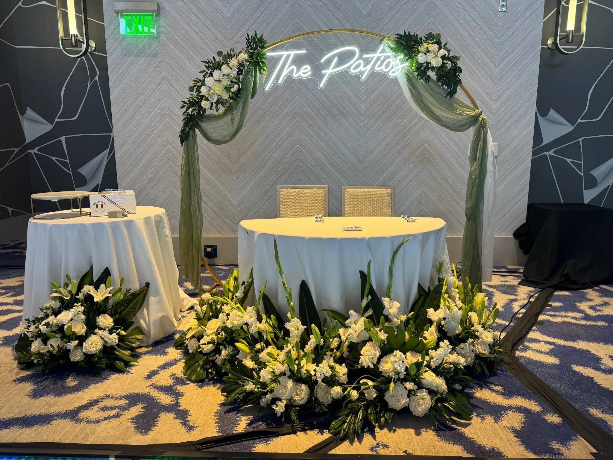 Decorative setup with a floral arch and flowers on the ground, with a table on the left and a backdrop with neon lights spelling 'The Plac' in the center.